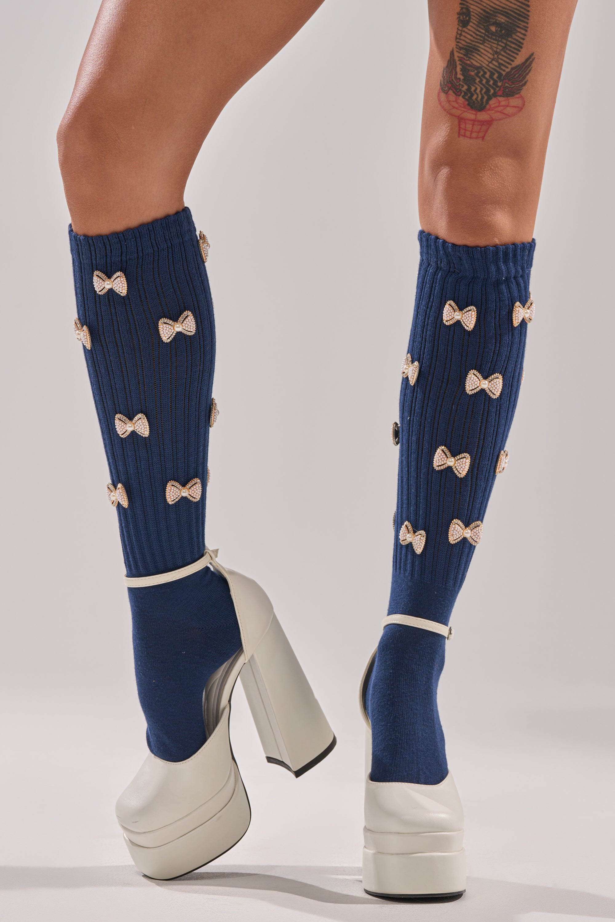 A person wears the BOW PLEASE CHARM SOCKS—navy, knee-high with small white bows—paired with white high platform heels. A colorful upper thigh tattoo adds flair to the look, set against a plain light gray background.
