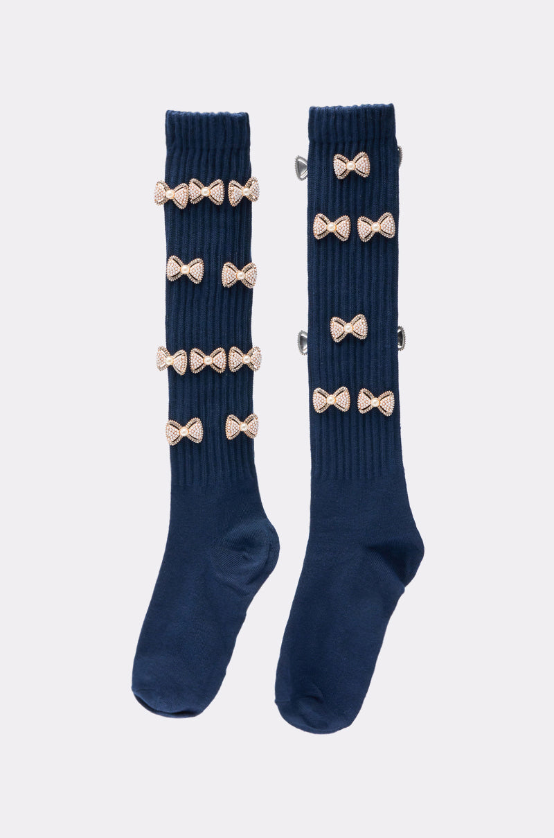 The BOW PLEASE CHARM SOCKS are navy knit socks, each featuring five gold glitter bows in two vertical columns down the front, displayed flat on a light grey background. 3