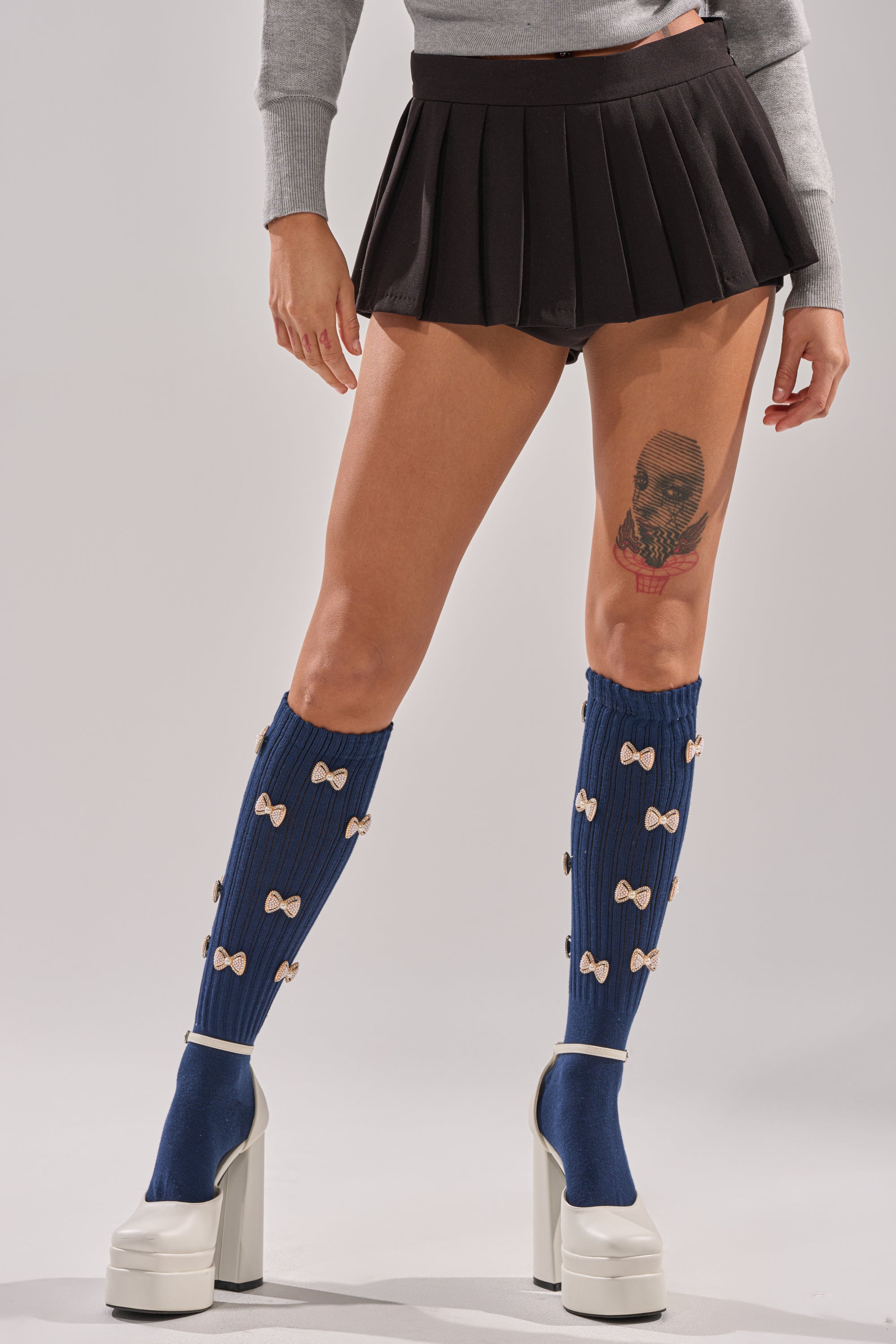 Wearing BOW PLEASE CHARM SOCKS—navy knit socks with bow decorations—paired with a short black pleated skirt, white platform heels, and a grey sweater. The cropped image focuses on the lower body and shows a visible leg tattoo.