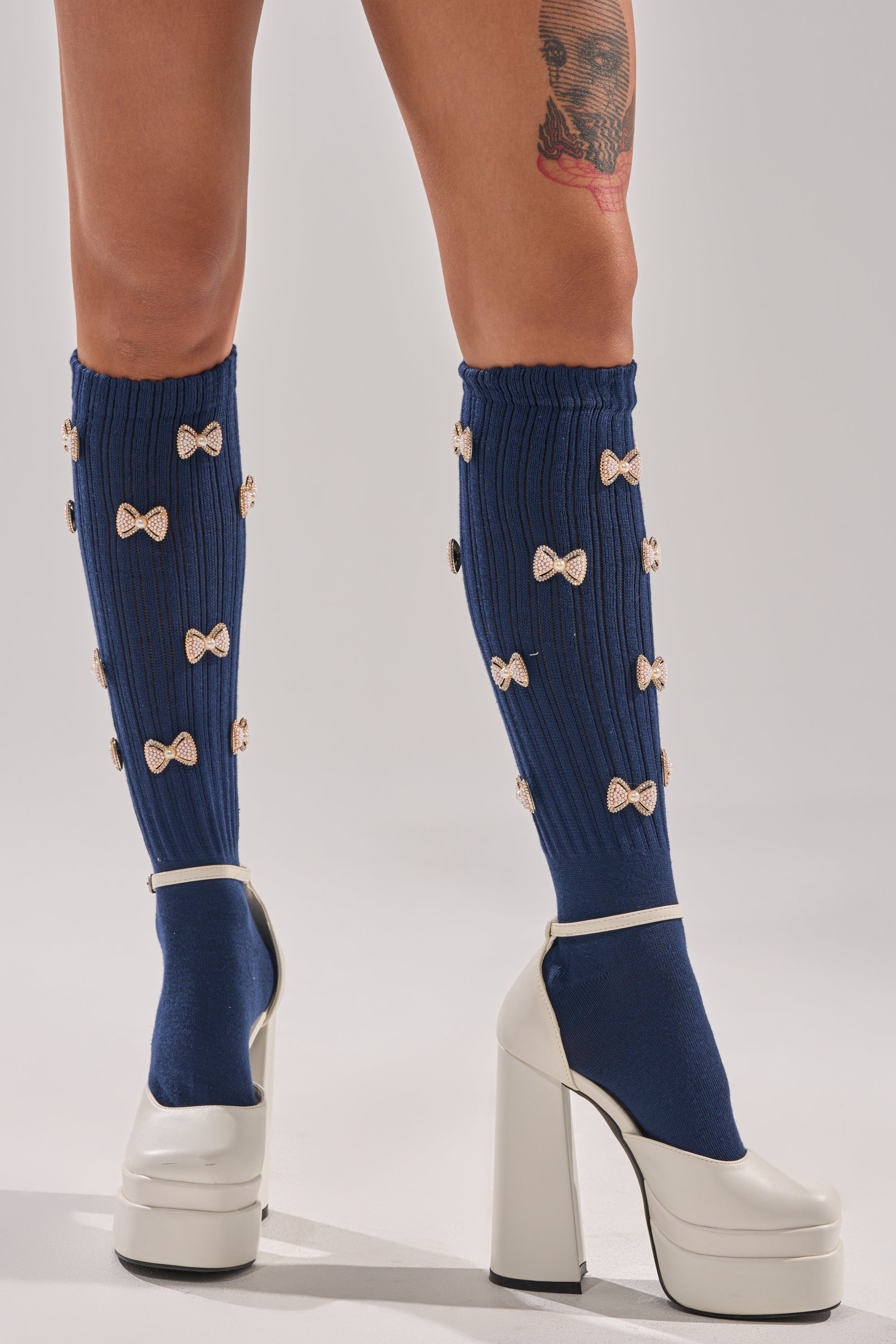 A person wears BOW PLEASE CHARM SOCKS—navy knit socks with small bows—and white chunky high-heeled platform shoes. A tattoo appears on their upper thigh.