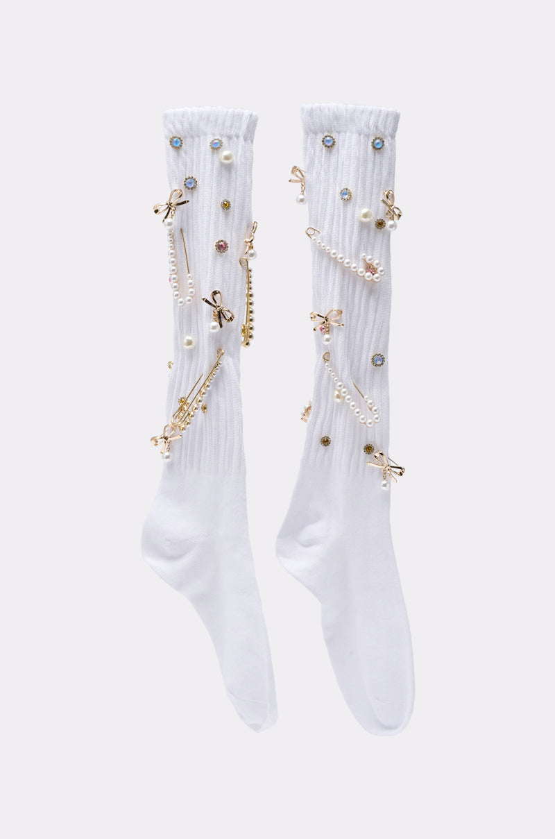 The ON TREND CHARM SOCKS are long white socks adorned with pearls, gold bows, chains, and colorful gem-like details, shown against a simple light background. 2