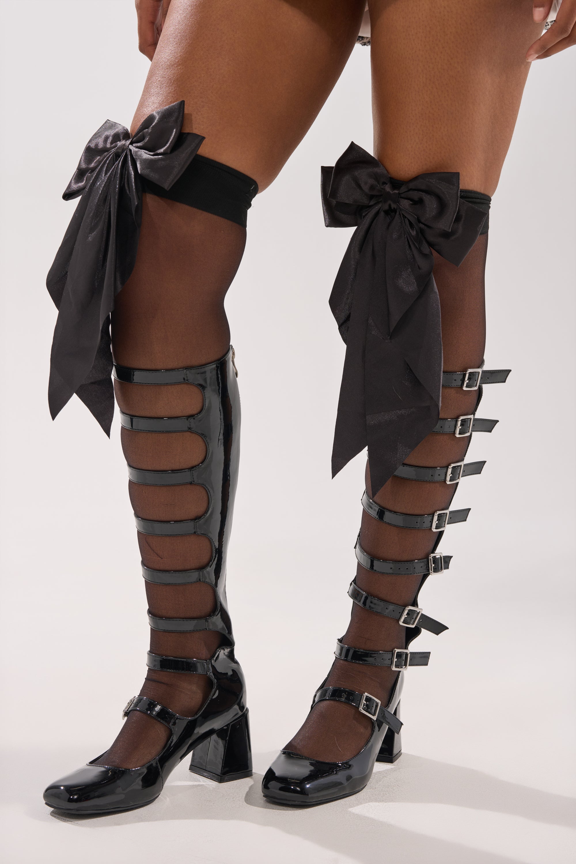 Someone wearing DRESS ME UP BOW STOCKINGS—sheer black stockings with large satin bows—paired with black patent gladiator-style knee-high boots featuring chunky heels and multiple buckle straps. 1