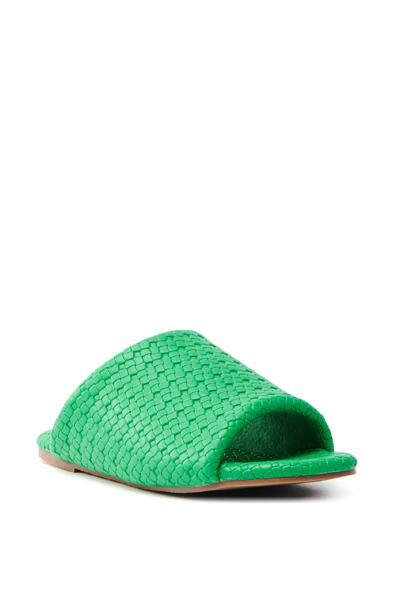 A close-up of the AKIRA GAIA Green Braided Flat Slip-On Sandal on a white background showcases its open-toed design. The upper features an intricate basket-weave pattern in vibrant green, complemented by a matching faux leather flat sole.