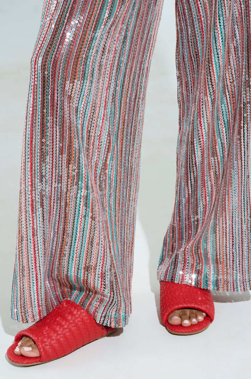The person is wearing colorful, sequined striped pants with vertical white, red, blue, and green stripes, perfectly paired with the AKIRA Gaia Red Braided Flat Slip On Sandals.