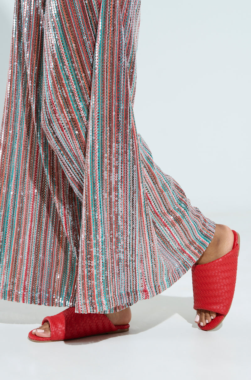 The image shows someone walking in wide-legged pants with red, green, and silver stripes on a light background, paired with the AKIRA GAIA RED BRAIDED FLAT SLIP ON SANDALS.
