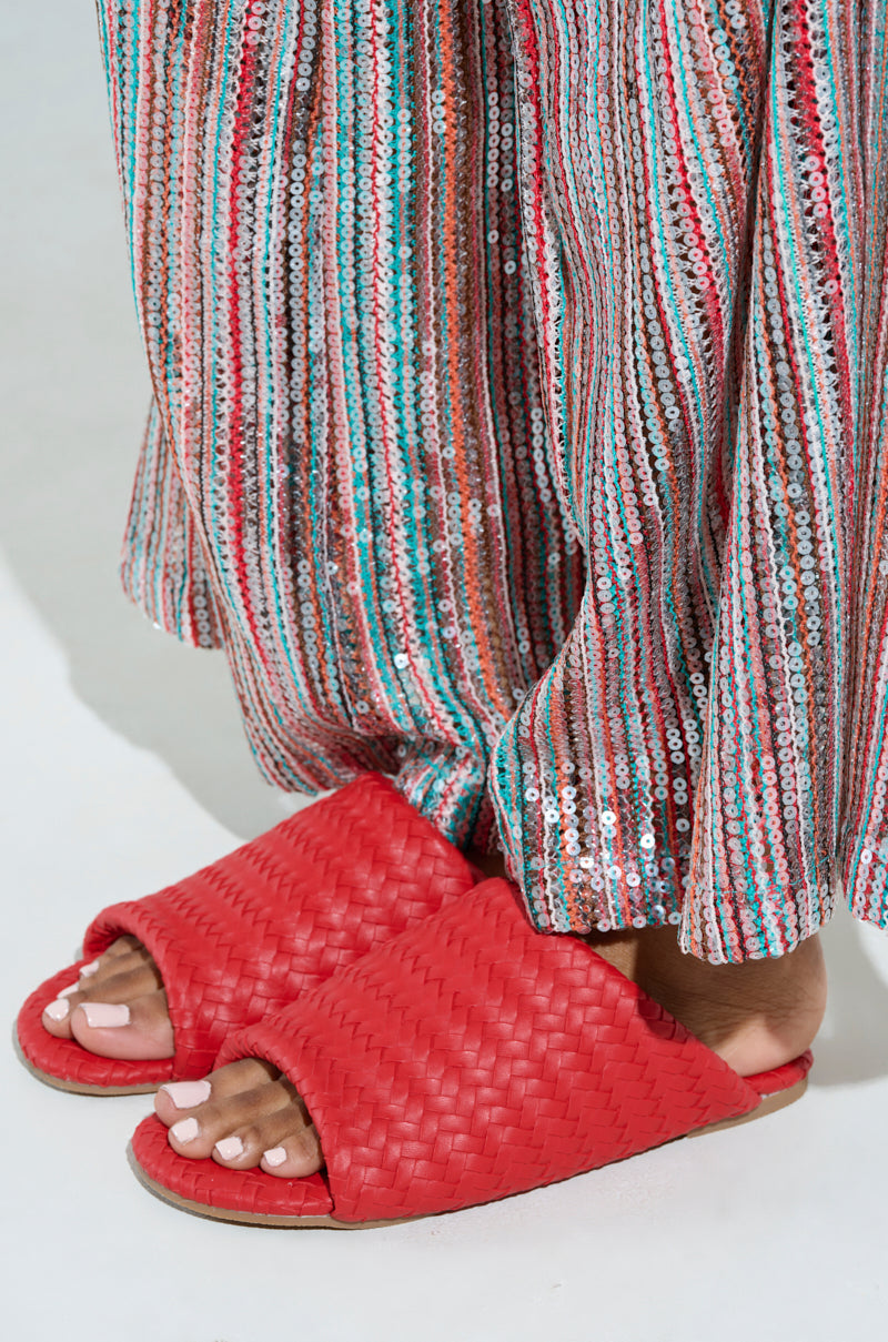 A person is wearing the AKIRA GAIA red braided flat slip-on sandals with neatly pedicured white toenails. Their long sequined skirt has vertical stripes of red, blue, and silver that catch the light beautifully.