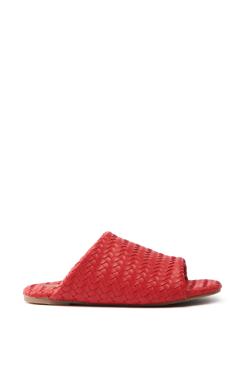The AKIRA GAIA RED BRAIDED FLAT SLIP ON SANDAL showcases a faux leather design with a cherry red braided upper, flat sole, and open toe, blending comfort and style against a white background.
