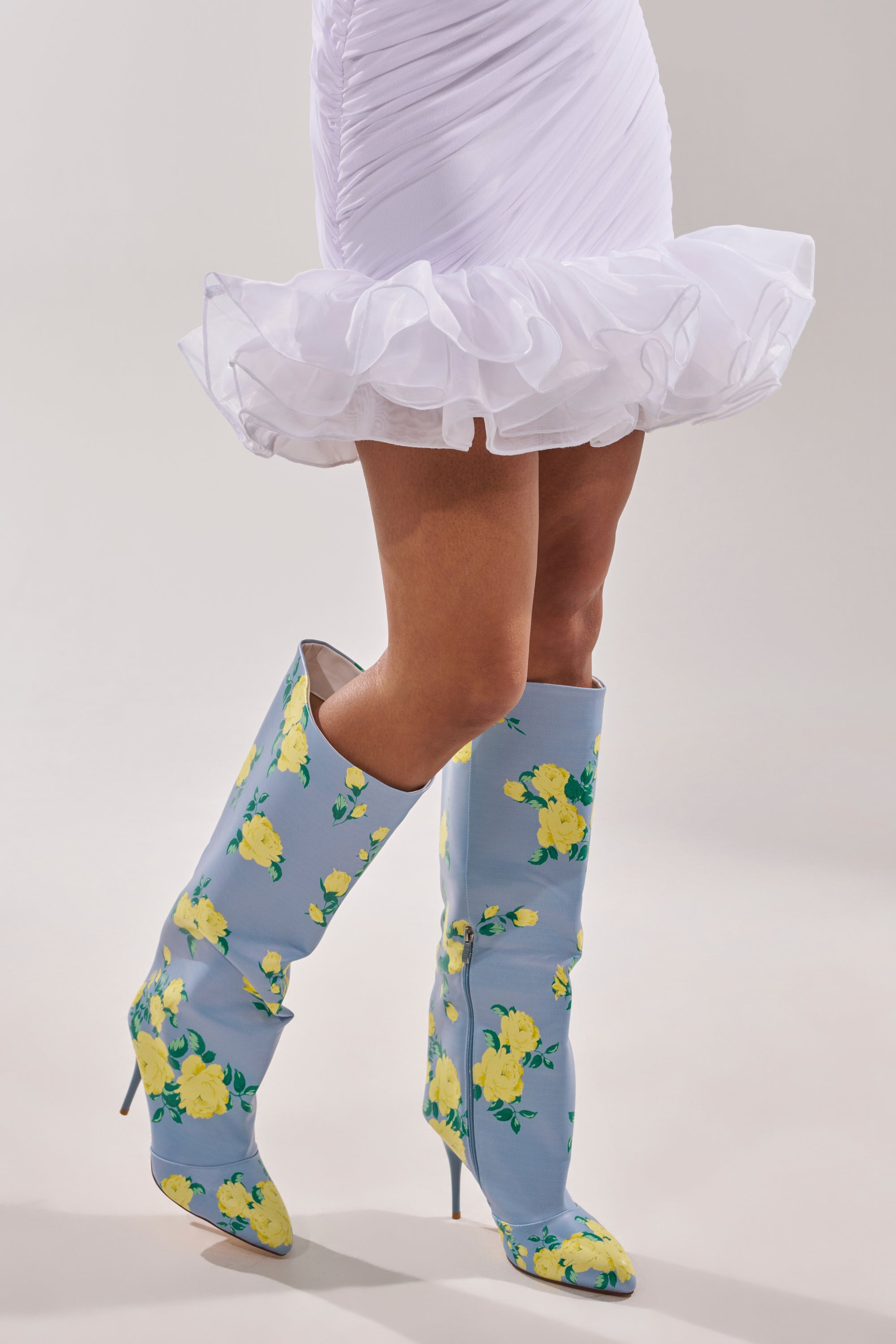 A person wearing a short, ruffled white dress and the AKIRA GOLD GARLAND BLUE FLORAL BOOT stands against a plain background. 1