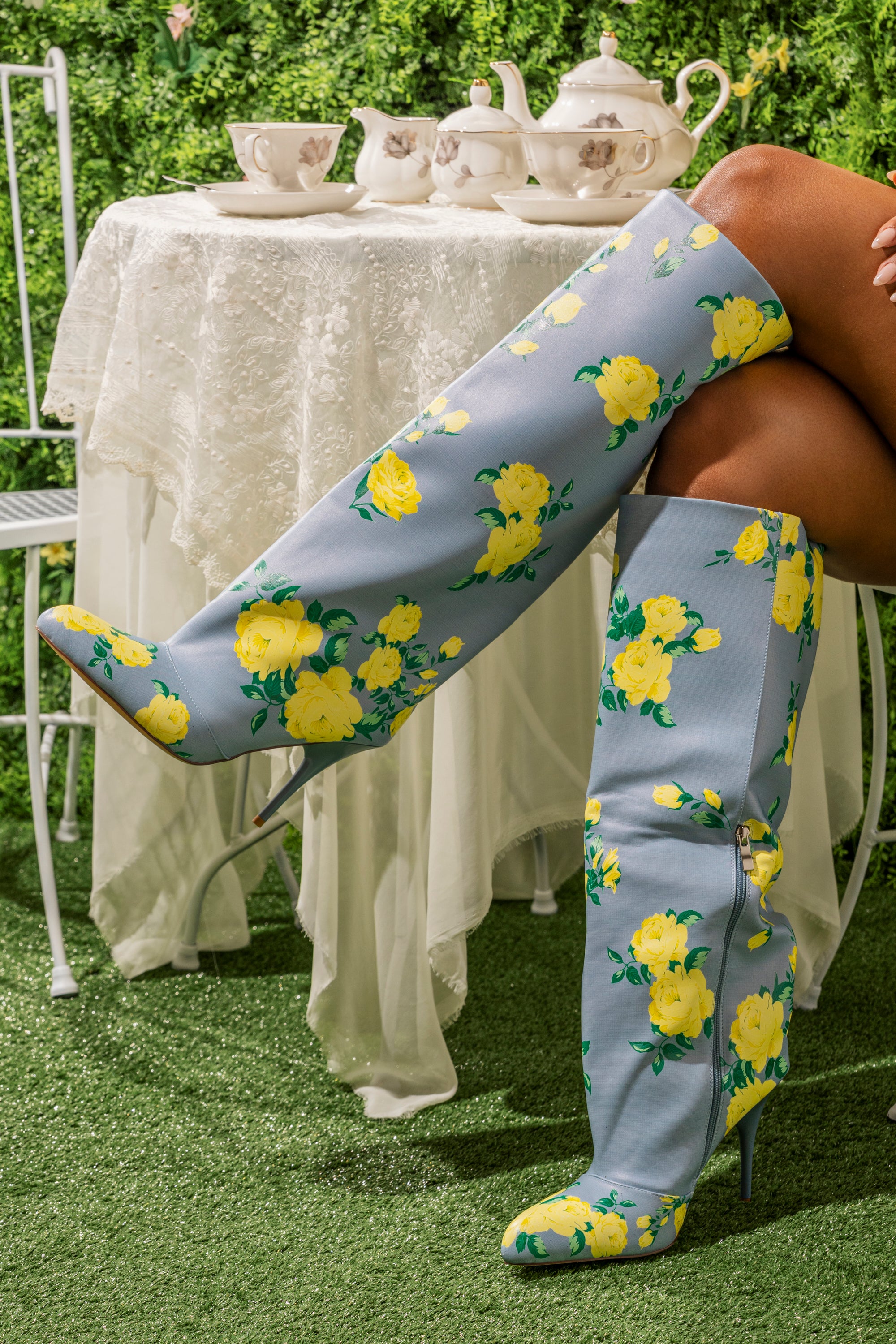 A person sits at a garden table wearing AKIRA GOLD GARLAND BLUE FLORAL BOOTS. Bright yellow flowers decorate the blue faux leather. A teapot and teacups rest on the lace-covered table in the background. 0