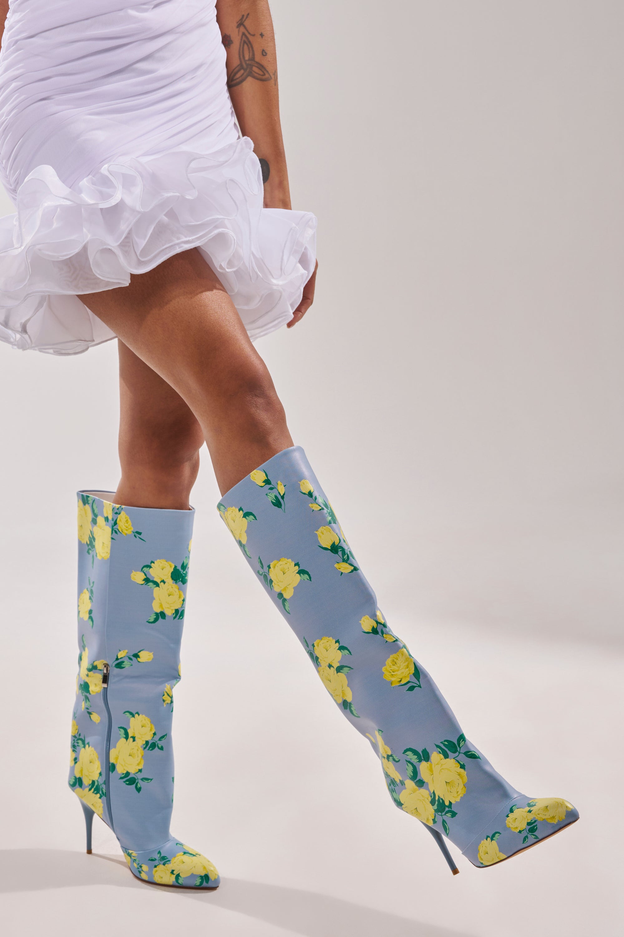 A person wearing a short, ruffled white dress poses with one foot pointed forward, showcasing the AKIRA GOLD GARLAND BLUE FLORAL BOOT against a plain background. 2