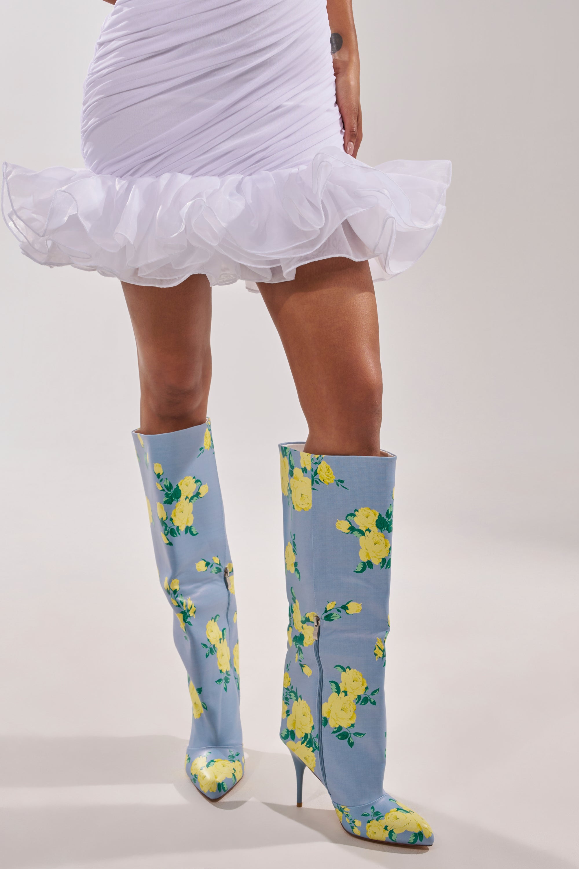 A person wears a white ruffled mini dress and AKIRA GOLD GARLAND BLUE FLORAL BOOTs, set against a plain light background with a blue backdrop featuring subtle yellow floral accents. 4