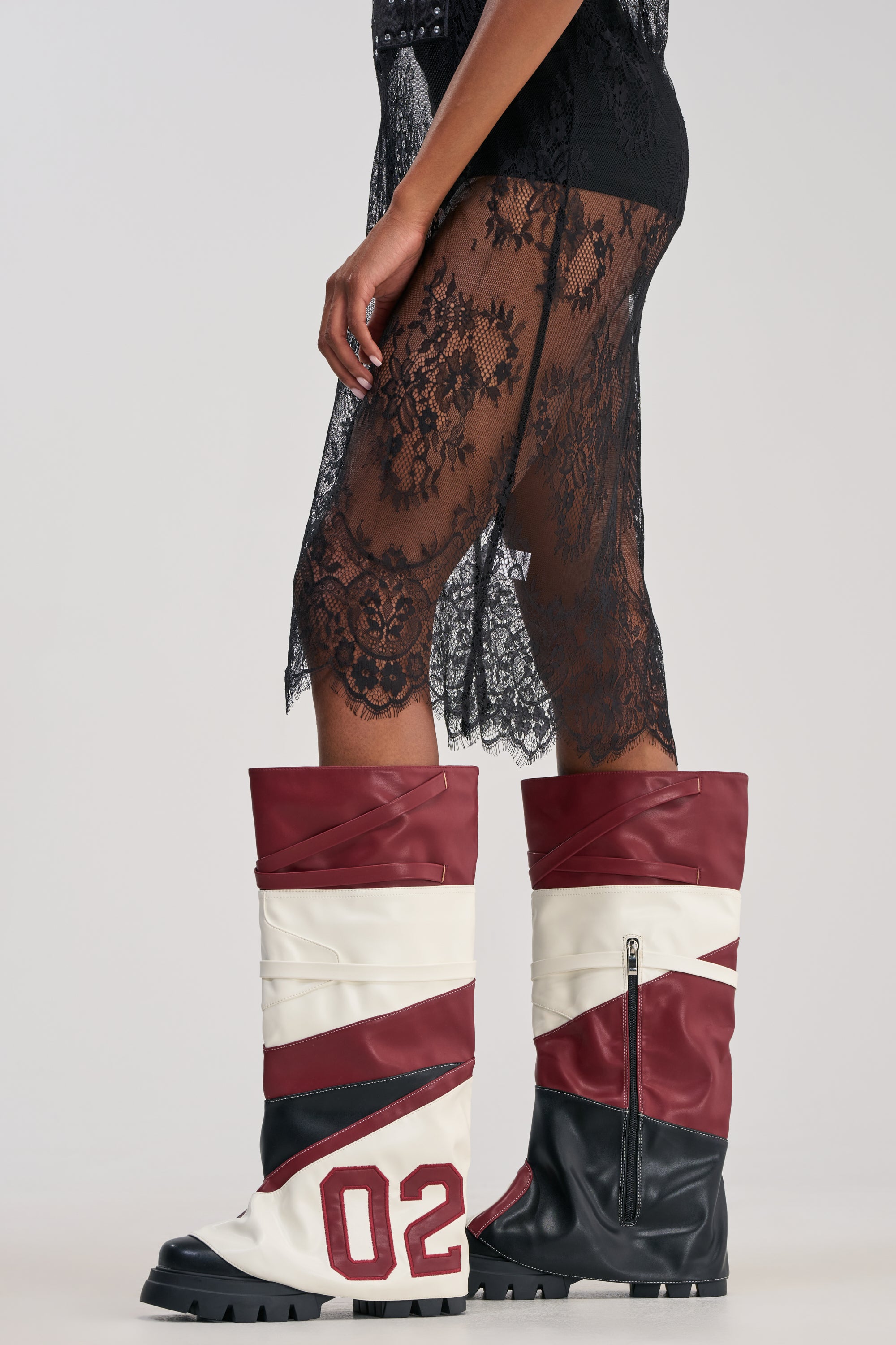 Wearing the AKIRA GOLD GARRICK Colorblocked Sport Combat Boot—a tall knee-high style in maroon, white, and black with a bold “02” on the side—paired with a black lace skirt, this look channels varsity streetwear vibes against a plain background.