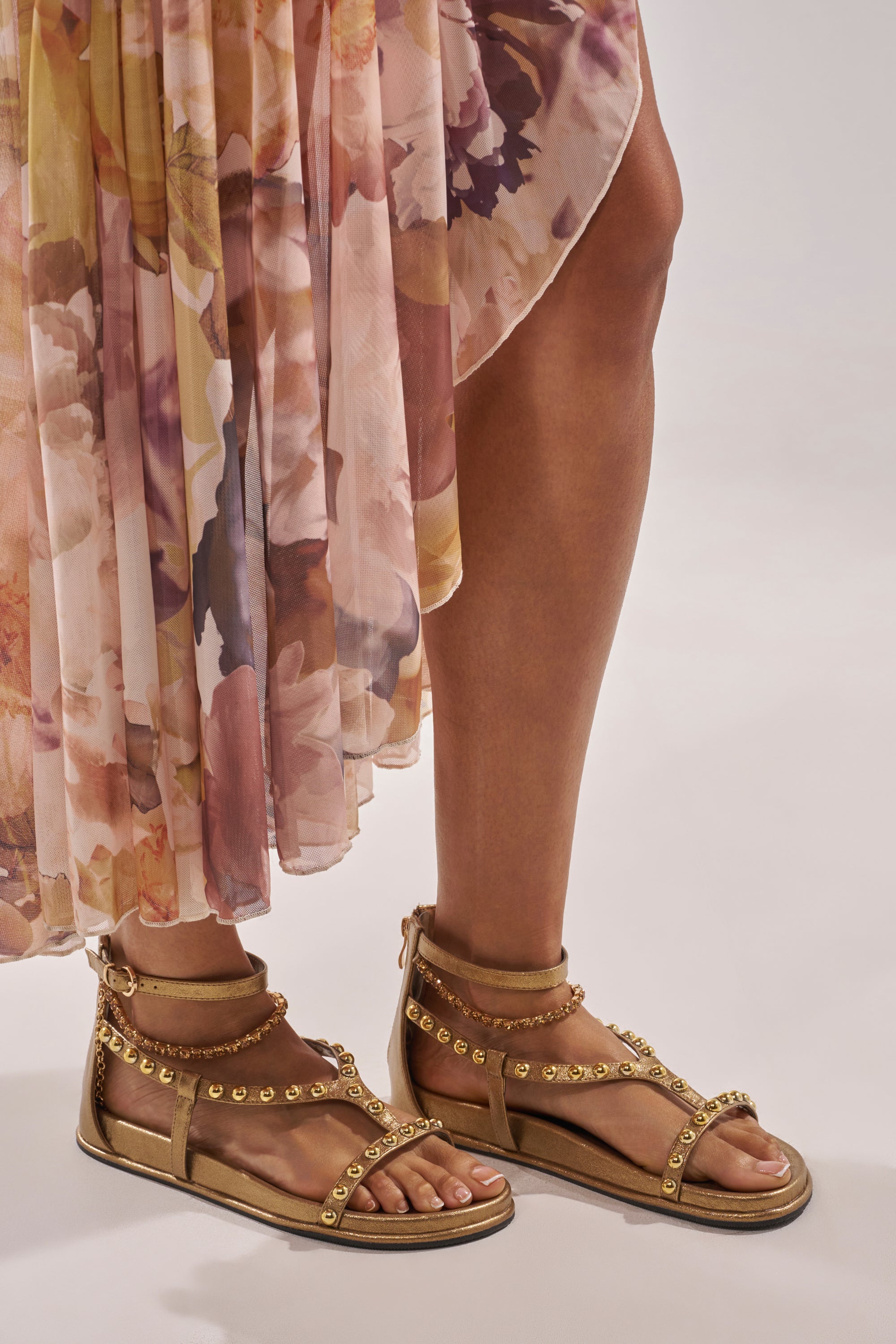 A person wearing a flowing, floral-patterned skirt and AKIRA GATO BRONZE GOLD METAL EMBELLISHED FLAT SANDALS stands on a light-colored surface. 0