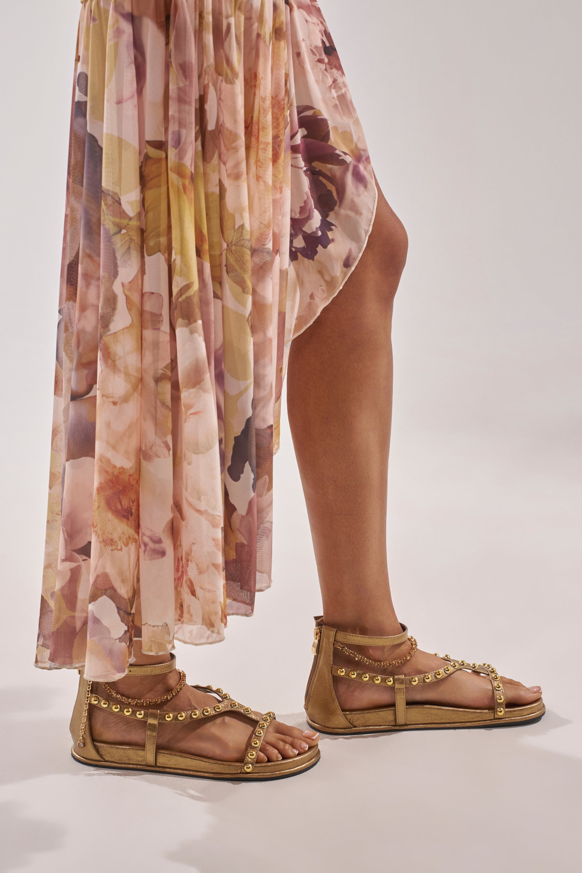 A person wearing a flowy, floral-patterned skirt and AKIRA GATO BRONZE GOLD METAL EMBELLISHED FLAT SANDALS stands against a light, neutral background. 1