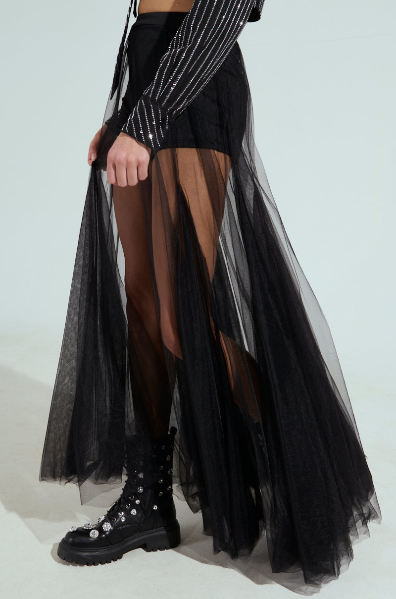 Someone is dressed in the MY ALL MAXI TULLE SKIRT, a sheer black maxi layer over black shorts, paired with a striped long-sleeve top. The outfit showcases an elegant high-rise style and is complemented by black boots adorned with silver details against a plain light grey background.