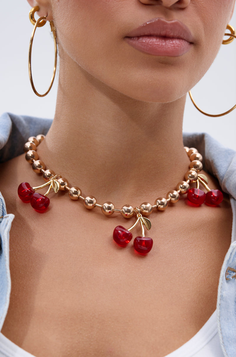 A woman in a light blue collared shirt is shown from the shoulders up, wearing large gold hoop earrings and the POP MINE NECKLACE—a gold-tone ball chain necklace with red cherry-shaped charms.