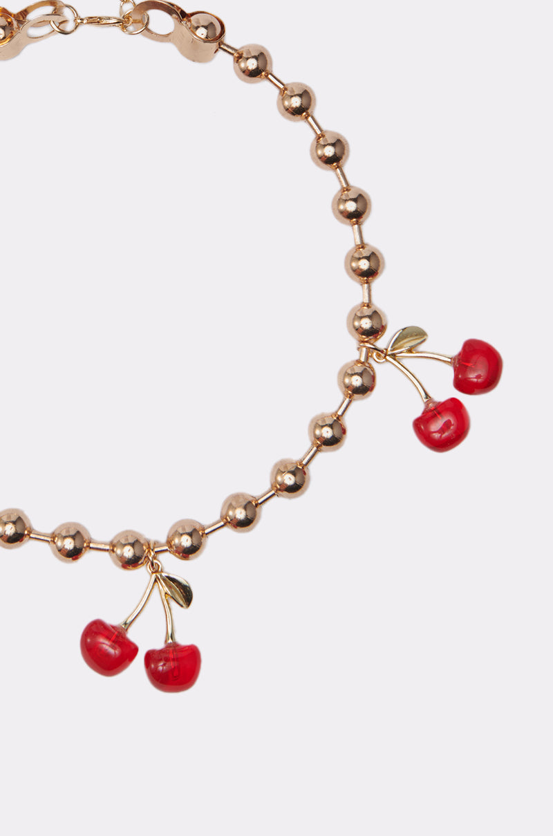 The POP MINE NECKLACE features a gold-tone ball chain with two cherry-shaped charms, each displaying red cherries and green stems, elegantly set against a plain white background.