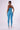 A woman is standing with her back turned, wearing the strapless CONSTANT LOVE JUMPSUIT by J.our in blue, featuring a bodycon fit and clear high-heeled shoes. She accessorizes with hoop earrings and sunglasses. The clean, simple setting features a white background.