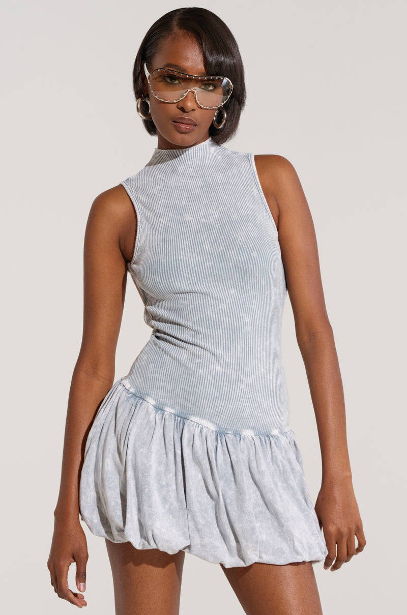 A woman with short, straight hair is wearing "THE BEST OF THE BEST MINI DRESS" from the AKIRA Label, which is a sleeveless, stretchy ribbed knit light blue dress featuring a high neckline and a short, ruffled skirt. She accessorizes with large round hoop earrings and wears oversized, transparent sunglasses. The background is plain and light.