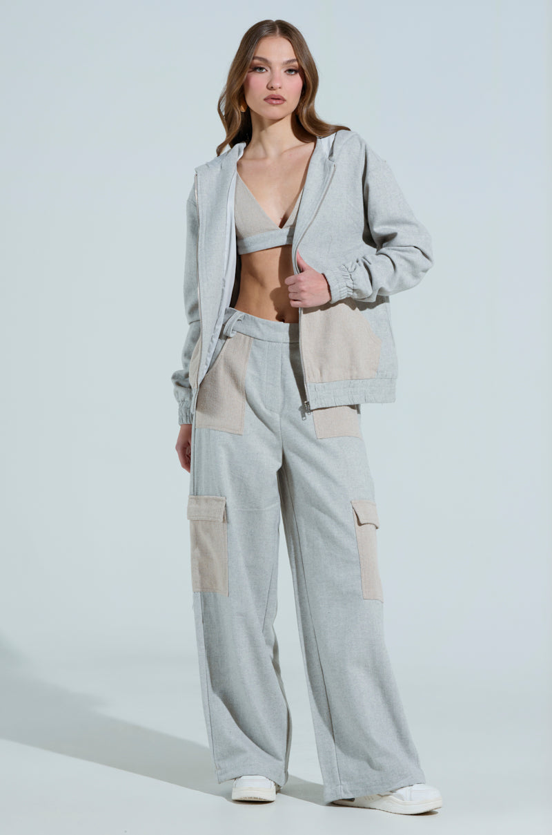 A person confidently wears TO LOVE AGAIN TROUSER with a high-rise waistband and wide-leg design. They pair it with a zipped hoodie over a matching crop top, and white sneakers, standing against a plain light gray background in a light gray and beige outfit.