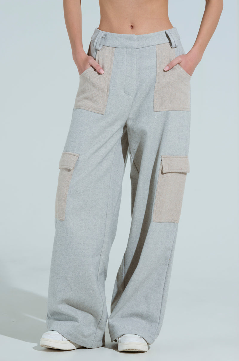Someone is wearing the TO LOVE AGAIN TROUSER from AKIRA Label, featuring light gray cargo pants with a high-rise waistband, wide leg silhouette, and beige fabric accents. They pair them with white sneakers against a plain, light-colored backdrop.