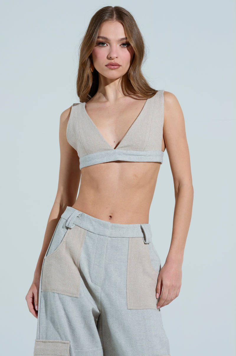 A person with long hair models the TO LOVE AGAIN BRALETTE in gray, featuring a plunging neckline. They pair it with high-waisted trousers with large pockets, standing against a plain light blue background and gazing directly at the camera.