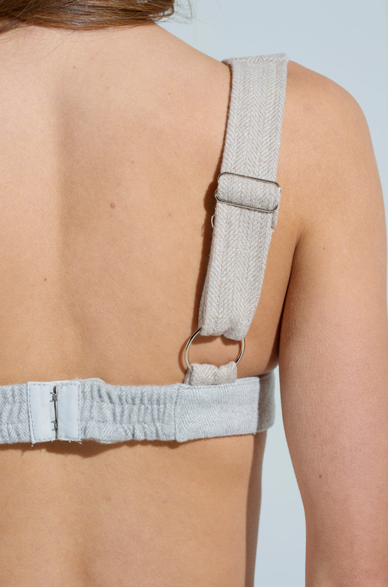 A close-up of a person's back highlights the TO LOVE AGAIN BRALETTE by AKIRA Label, showcasing its beige fabric strap with metal hardware. The strap crosses over the shoulder and fastens with a clasp at the back against a soft, light background.