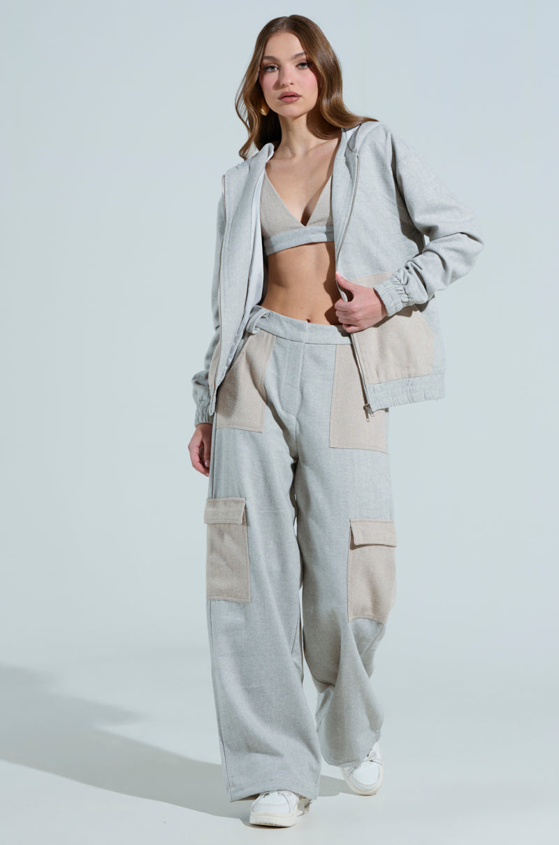 A woman confidently strides towards the camera, wearing a light gray tracksuit with large pockets and the TO LOVE AGAIN BRALETTE. Her long hair cascades elegantly as she maintains a neutral expression against a plain, well-lit background, embodying the AKIRA Label To Love Again aesthetic.