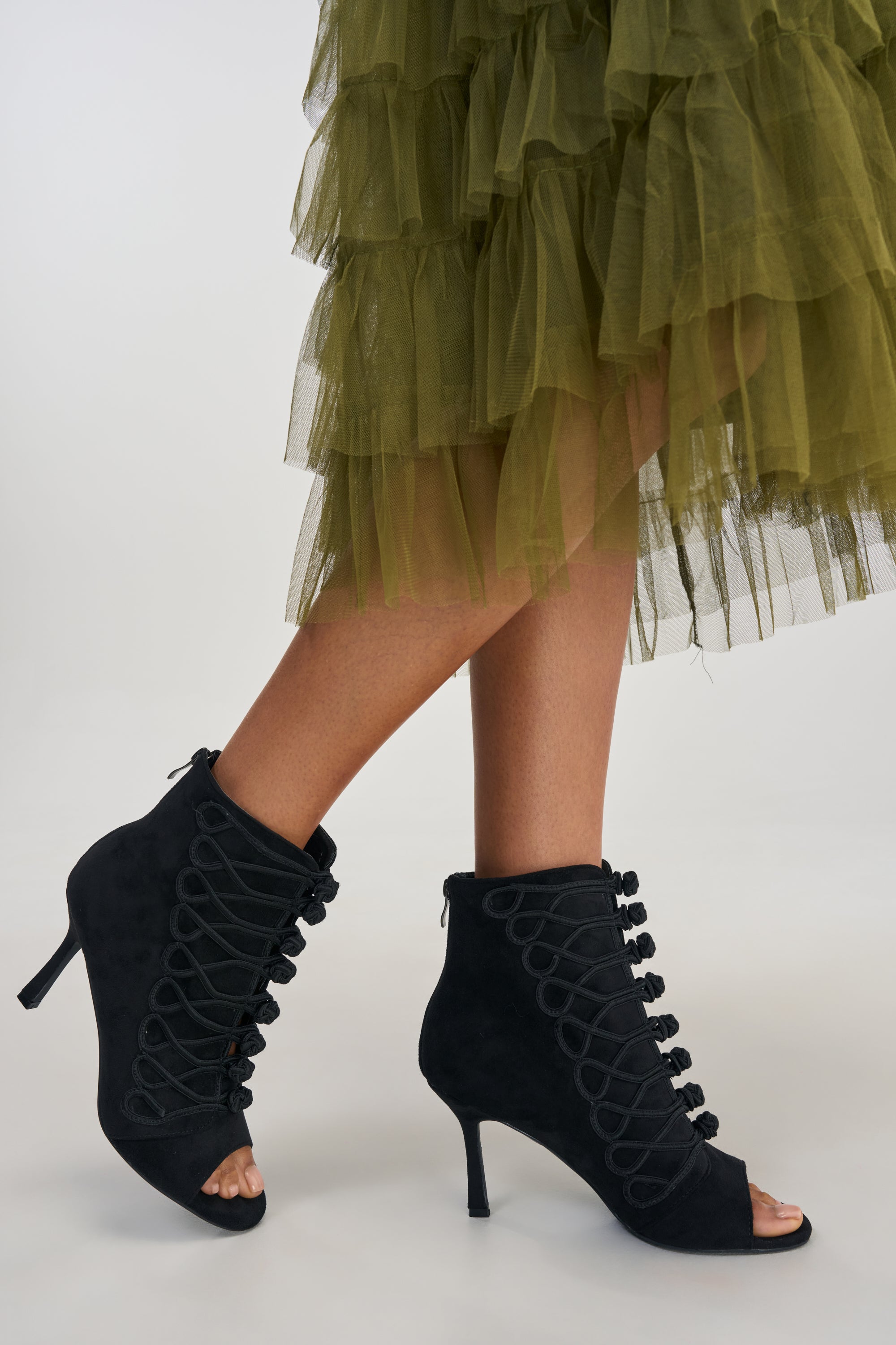 Wearing the AZALEA WANG GENTRY BLACK MILITARY STILETTO BOOTIE, a person stands on a white background, with a green ruffled tulle skirt visible from the knees down.