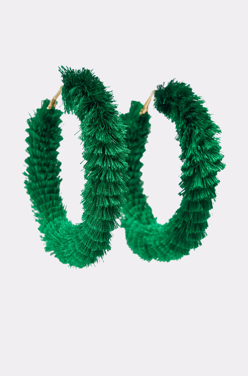 The CARNIVALE EARRING is an oversized, green fringe hoop earring with a thick, fuzzy texture, resembling artificial grass or pipe cleaners, shown against a plain white background. 2