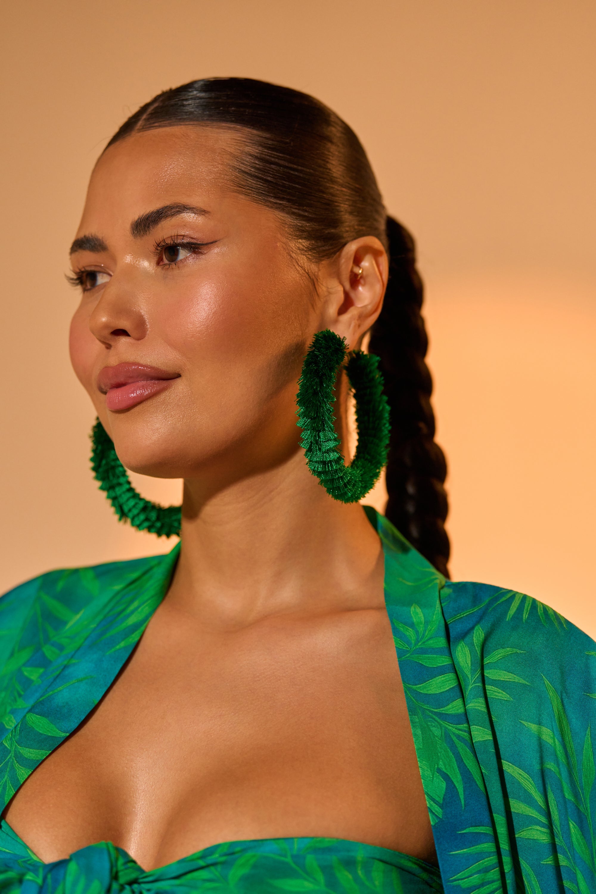 A woman with long dark hair in a braid wears the CARNIVALE EARRING—statement green fringe hoops—and a green leaf-patterned outfit. Her natural, glowing makeup and confident gaze embody festival jewelry style against a warm background. 0