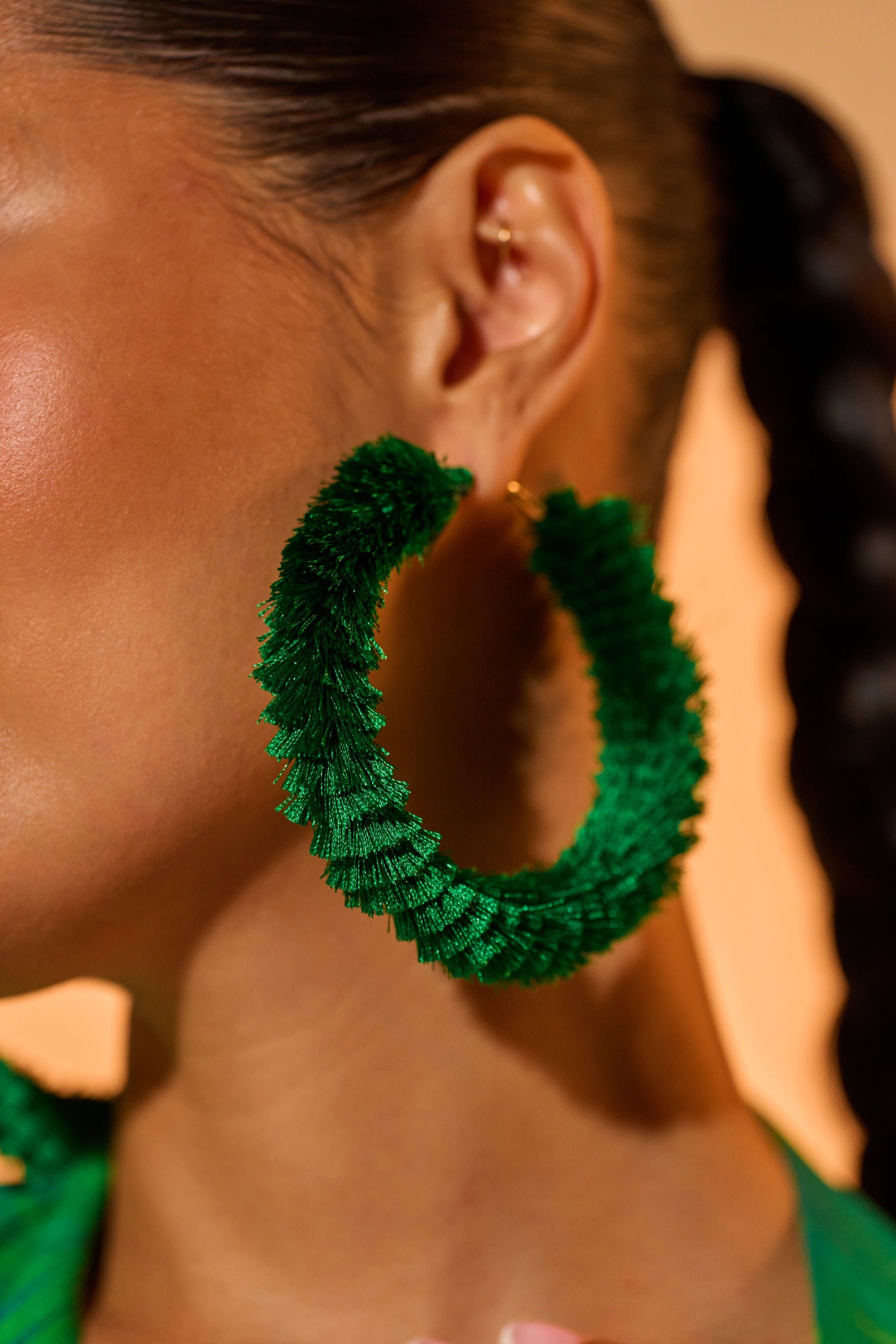 Close-up of a person wearing the CARNIVALE EARRING—large green fringe hoops. Hair is pulled back tightly, revealing part of their face and the statement earring against a softly blurred background—perfect festival jewelry inspiration. 1