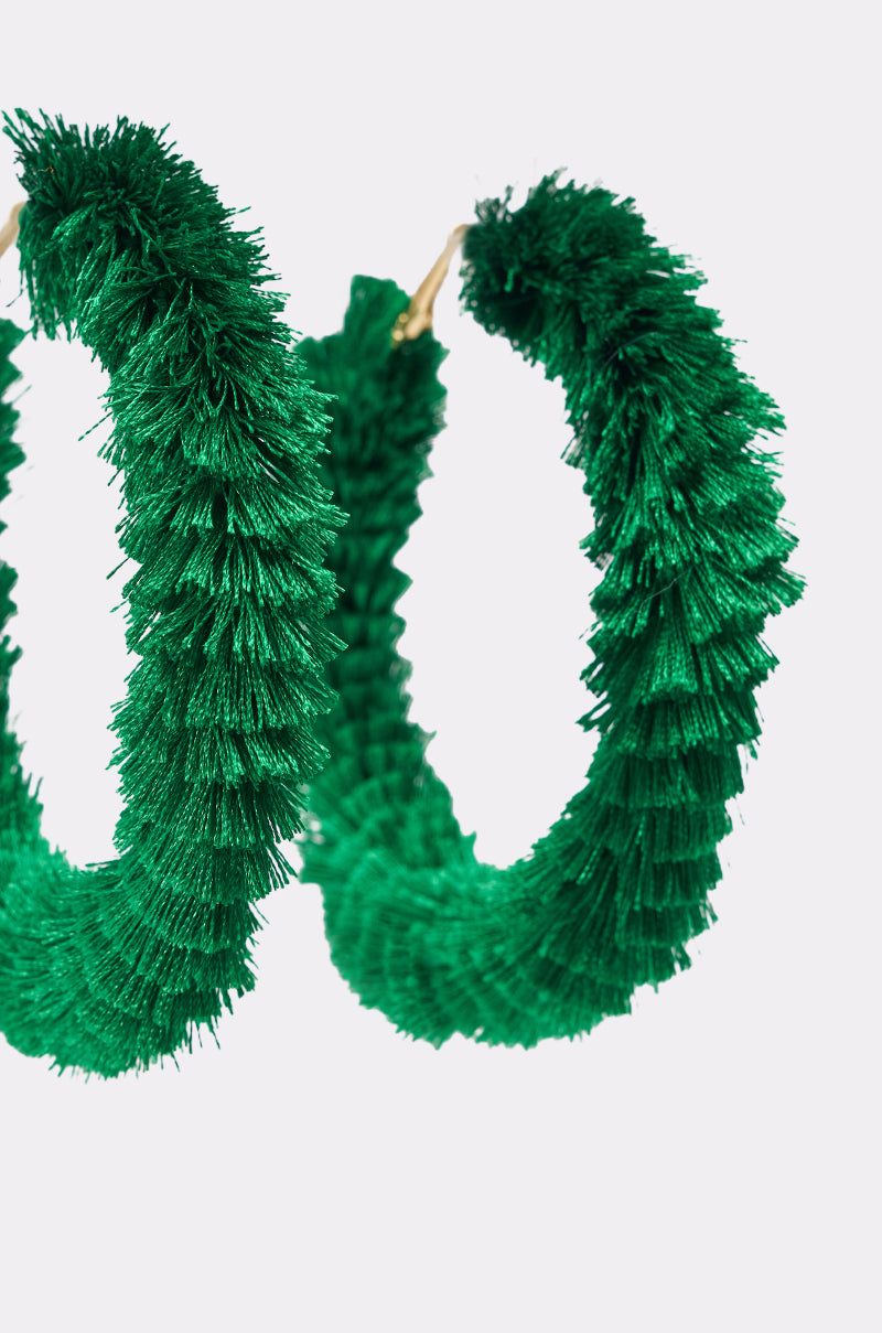 Close-up of the CARNIVALE EARRING, featuring two circular green hoops crafted from artificial pine-like material, resembling bold fashion earrings, set against a plain white background. 4