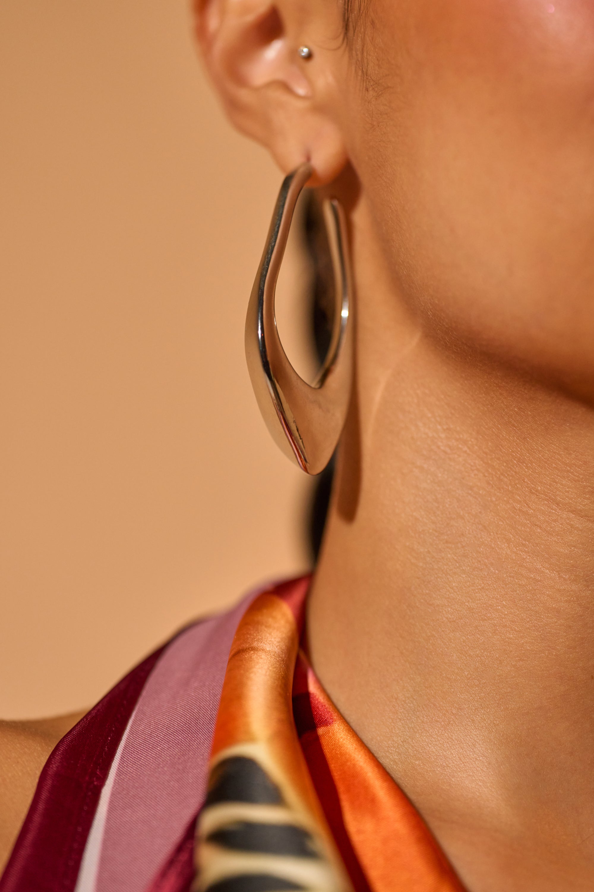 A close-up of a person's ear wearing the bold DRIPPY EARRING in an abstract silver-tone design, paired with a colorful scarf featuring red, orange, and black patterns. 0