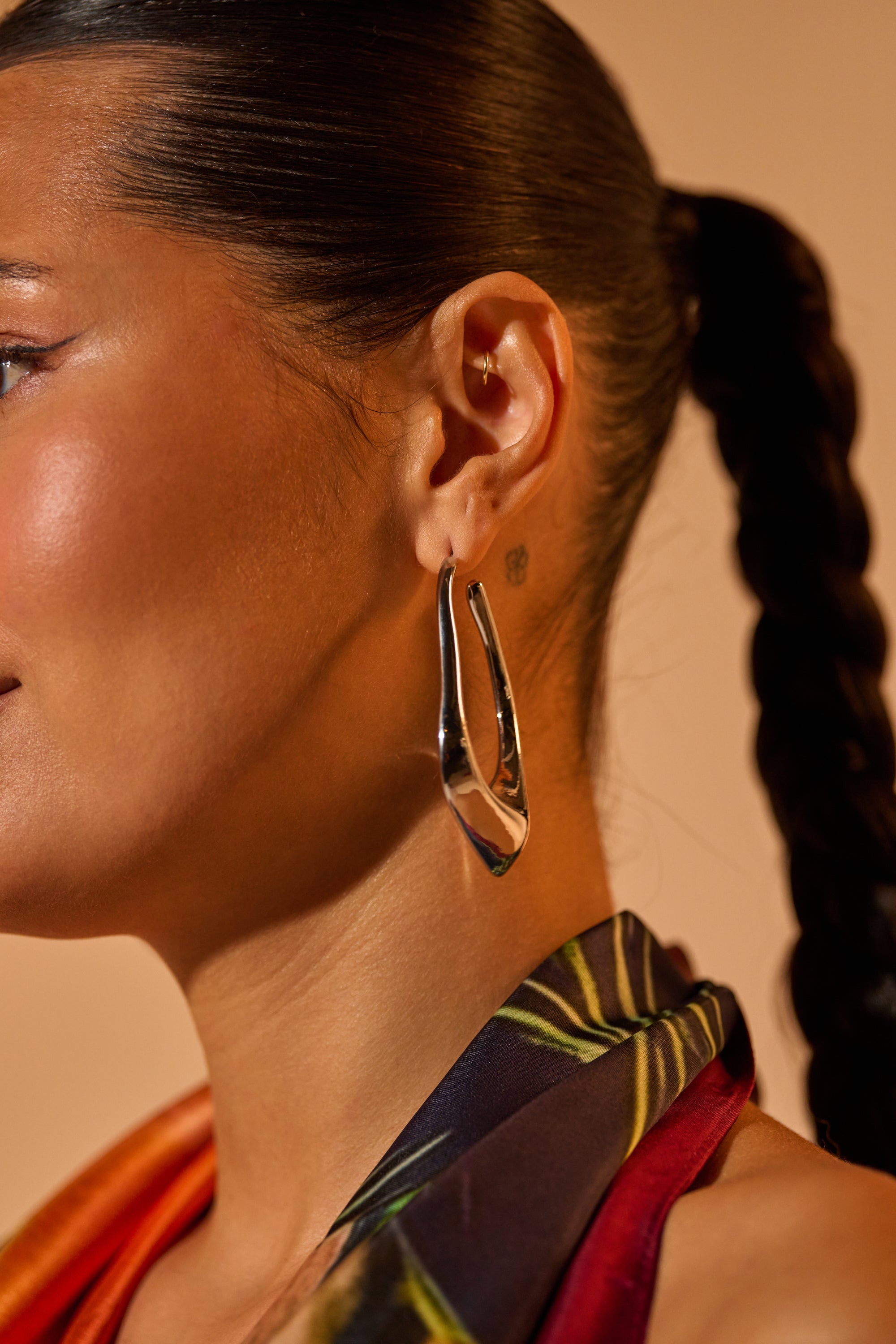 A close-up of a woman with a sleek, braided ponytail shows her wearing the DRIPPY EARRING in a silver-tone finish, paired with a colorful patterned top. 1
