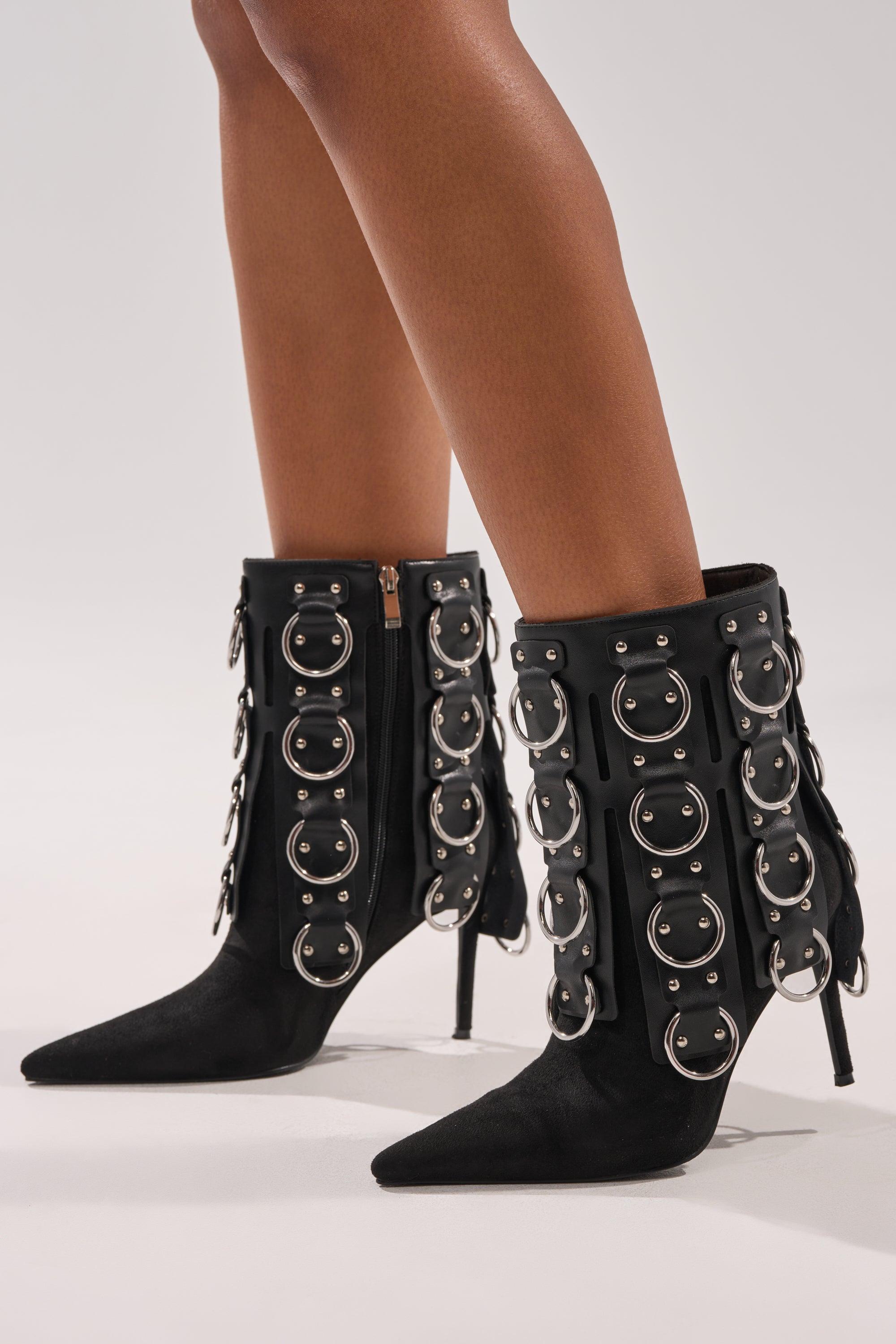 A person wears the AKIRA GOLD GERANIUM BLACK PIERCING STILETTO BOOTIE with circular metal rings, a sleek stiletto heel, and bare legs while standing on a light-colored surface.