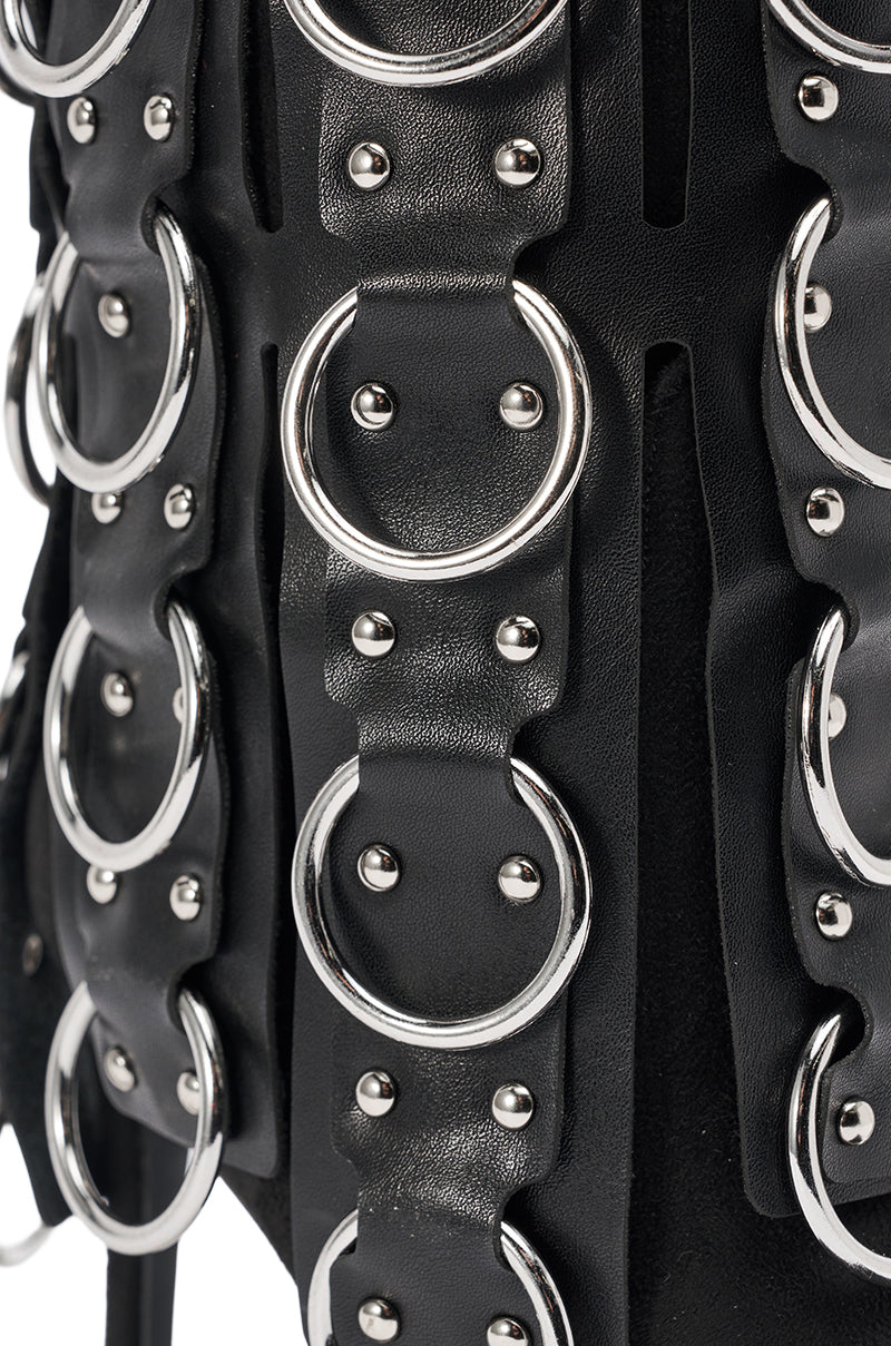 Close-up of the AKIRA GOLD GERANIUM BLACK PIERCING STILETTO BOOTIE, featuring black leather straps with large silver rings and rivets for a bold, textured look.