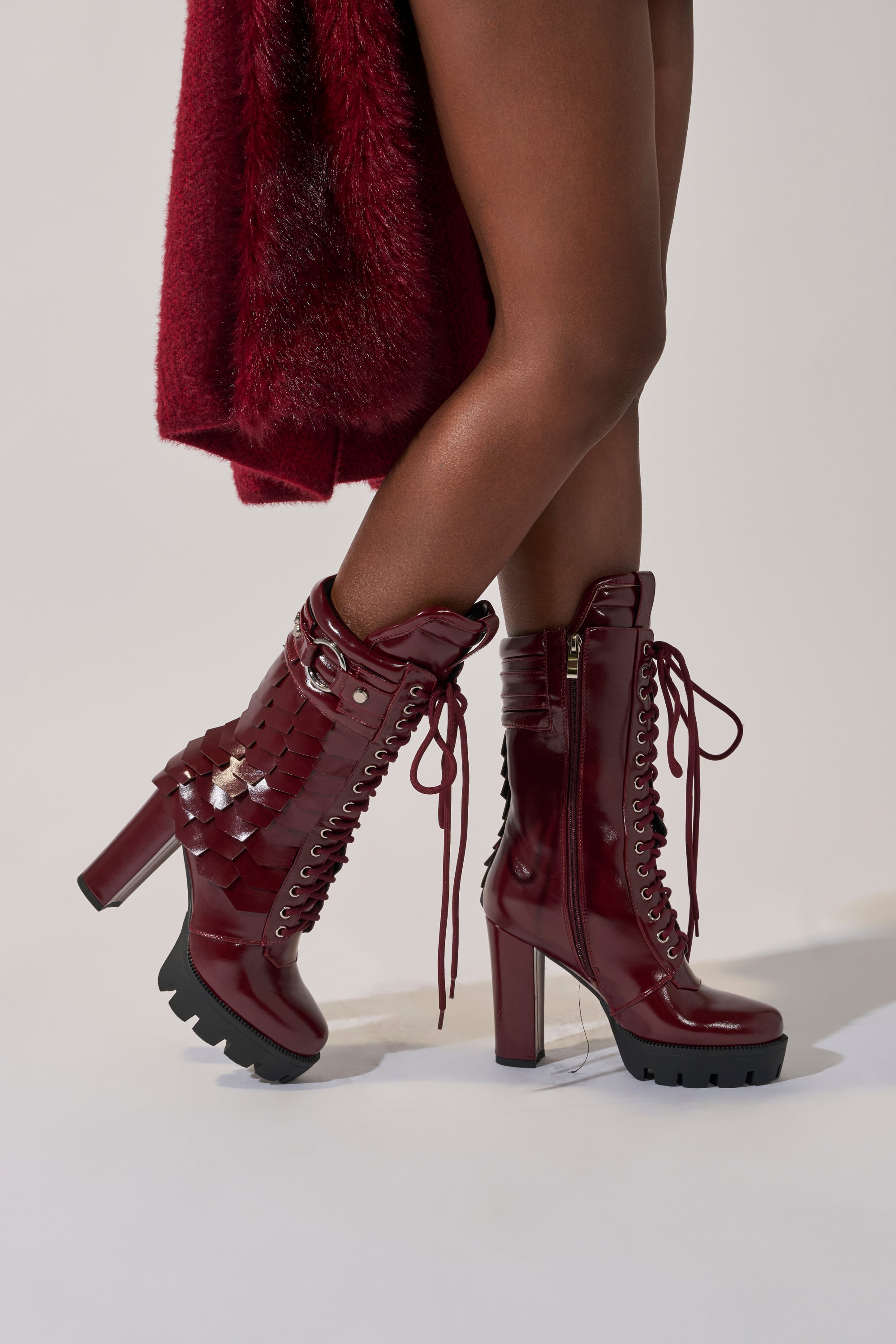 A person in a textured burgundy coat and the AZALEA WANG GERNOT Burgundy Lace Up Scaled Platform Bootie with chunky heels and metal details stands against a light background. 0