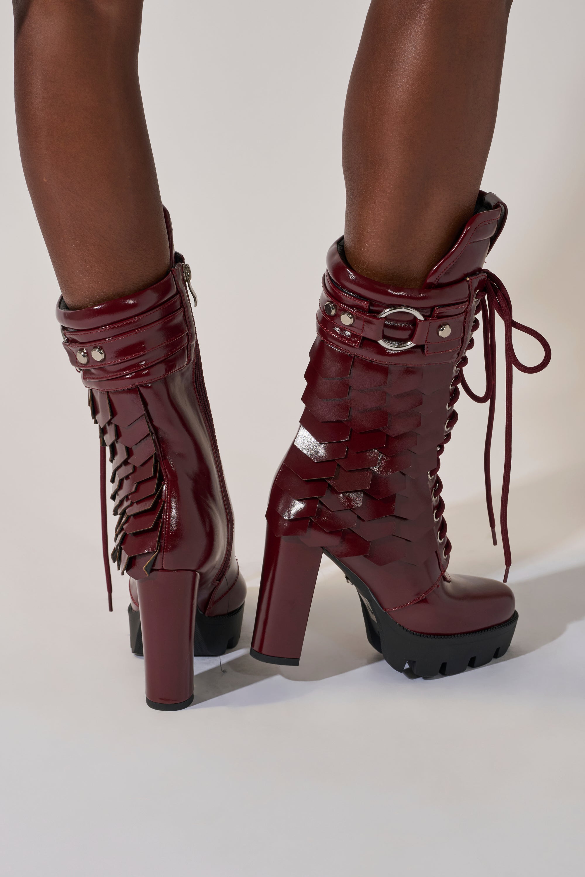 A person wears the AZALEA WANG GERNOT Burgundy Lace Up Scaled Platform Bootie, featuring chunky heels and bold details, standing against a light background. 2