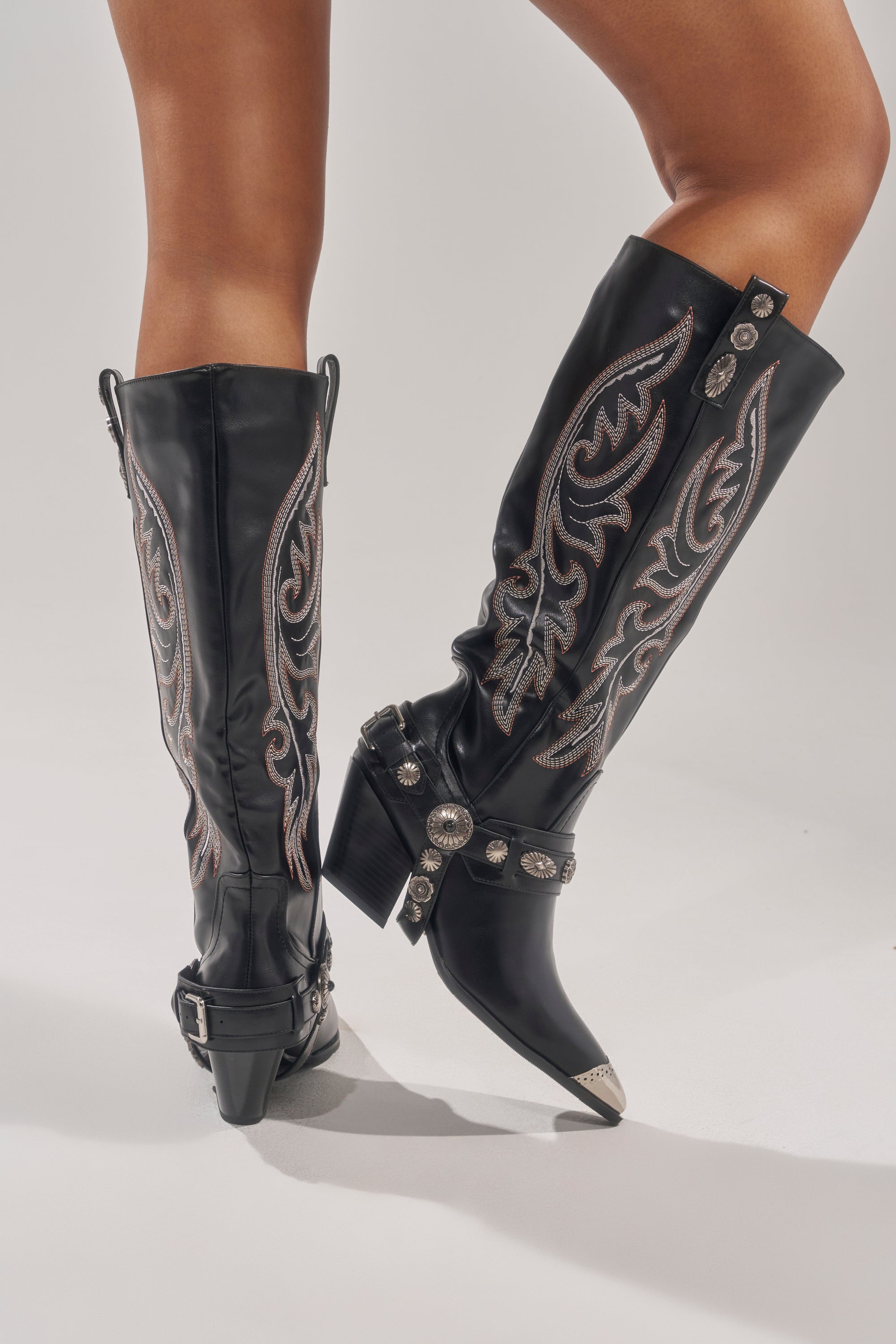 A person wearing the AZALEA WANG GERVASE BLACK PU SILVER HARDWARE WESTERN BOOT with silver hardware and harness-style straps stands on a light-colored surface. 0