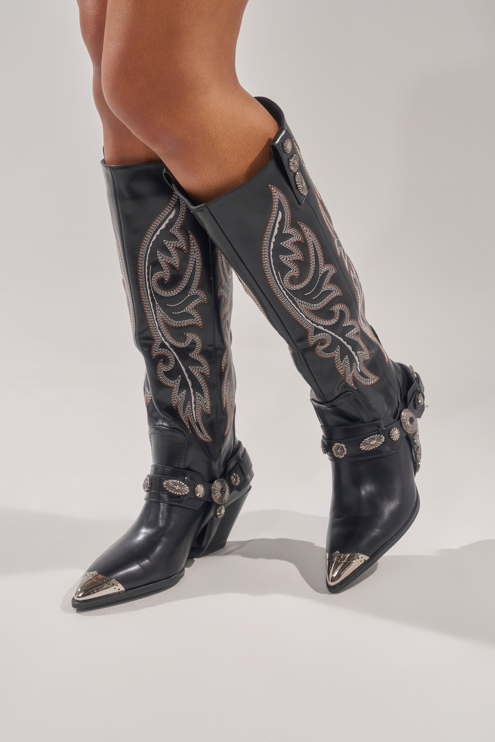 A person stands with legs crossed on a light gray background, wearing the AZALEA WANG GERVASE BLACK PU SILVER HARDWARE WESTERN BOOT featuring silver details and decorative straps. 1
