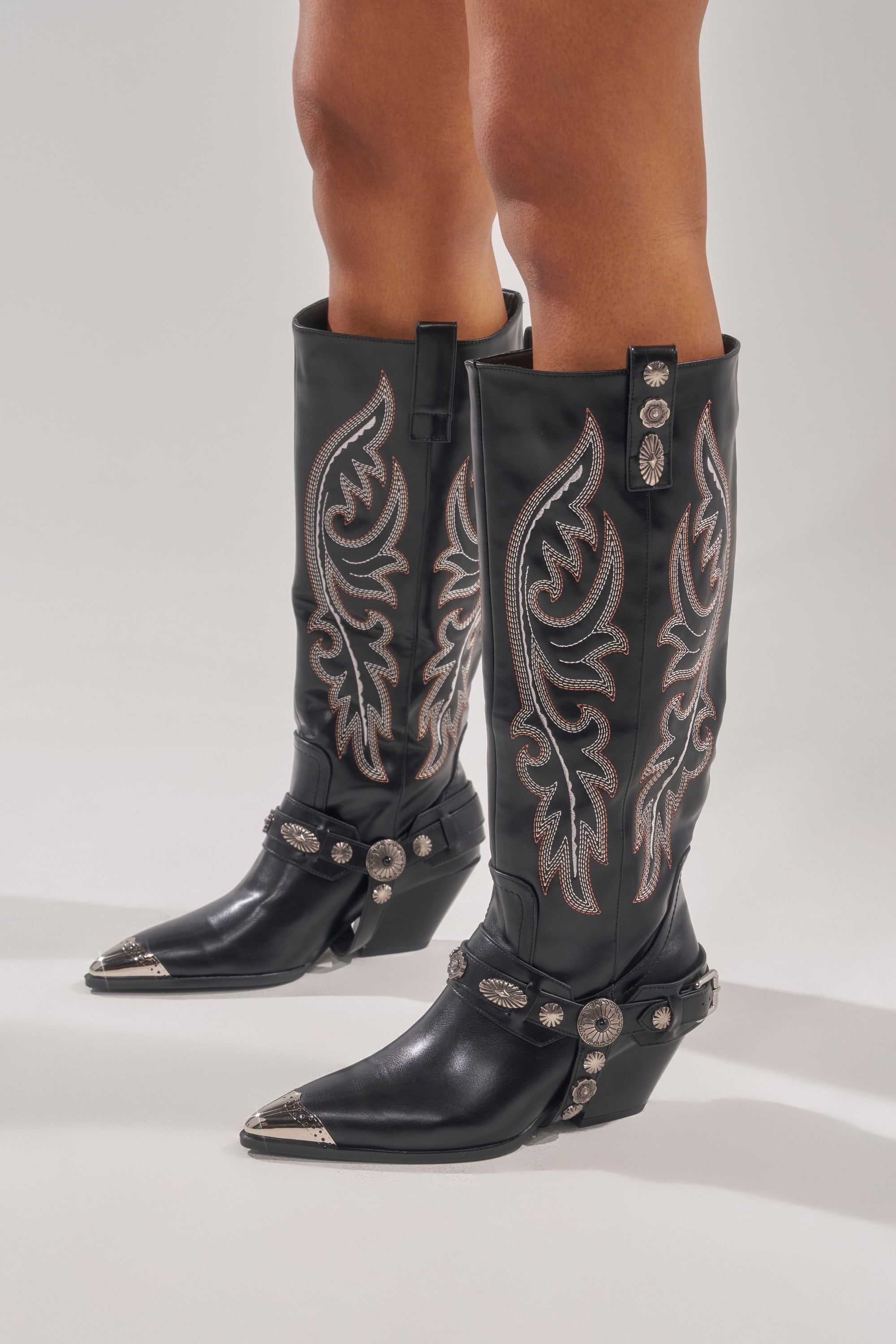 The AZALEA WANG GERVASE BLACK PU SILVER HARDWARE WESTERN BOOT features black PU, silver hardware, pointed metal-tipped toes, decorative straps, and chunky angled heels, shown on a person standing against a light background. 2