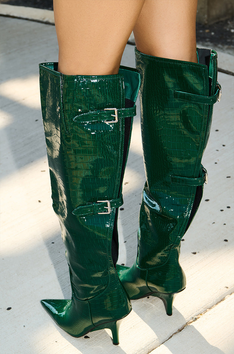 Someone stands on a light-colored sidewalk wearing the AZALEA WANG GESINE GREEN CROC PATENT LUXE BOOT—shiny, green, croc-embossed knee-high boots featuring pointed toes, stiletto heels, and decorative buckles.