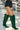 Wearing the AZALEA WANG GESINE GREEN CROC PATENT LUXE BOOT, black lace shorts, and a dark long-sleeve top, a person walks outdoors on a sunny day.