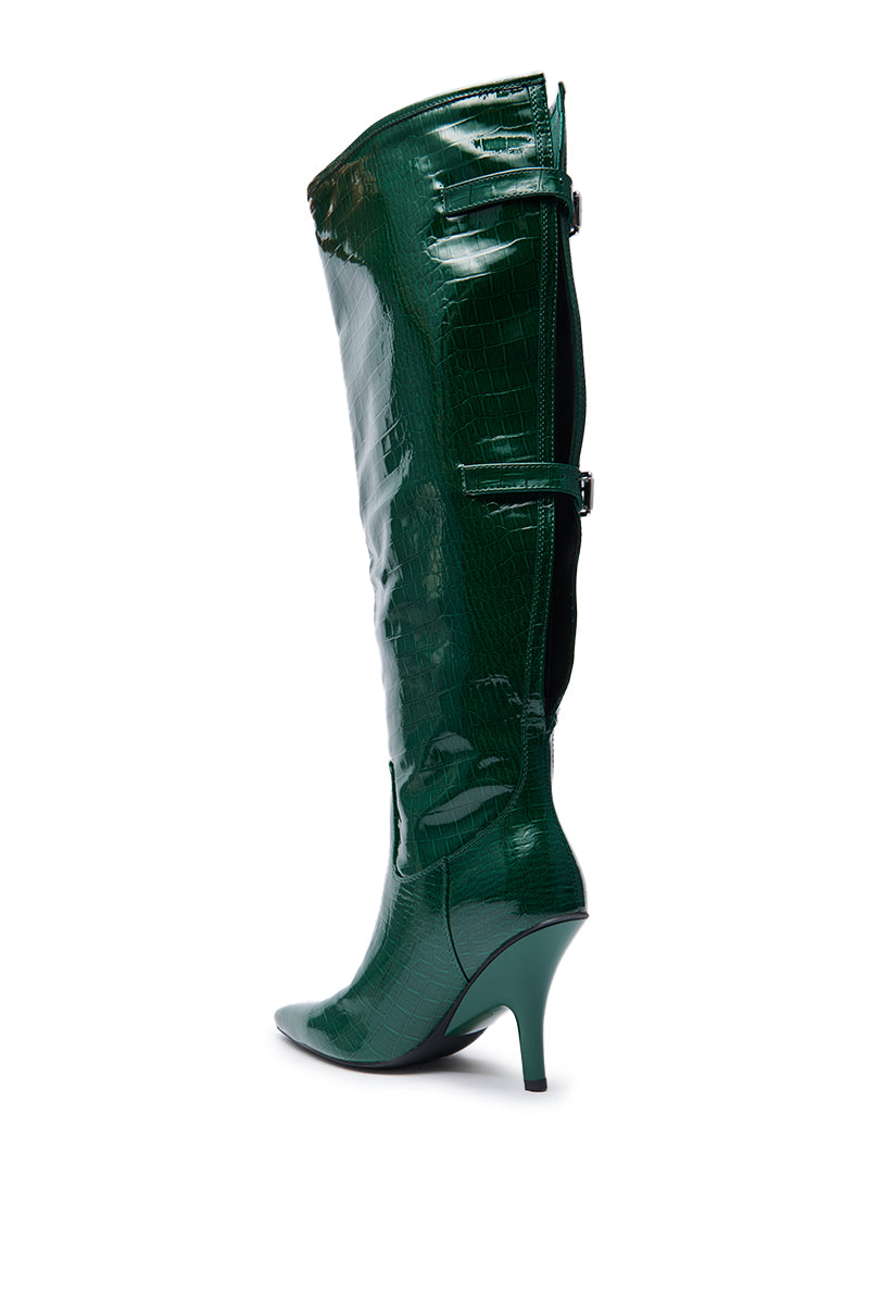 The AZALEA WANG GESINE GREEN CROC PATENT LUXE BOOT is a shiny, dark green, knee-high boot with a pointed toe, stiletto heel, croc-embossed pattern, decorative side straps, and a back zipper.