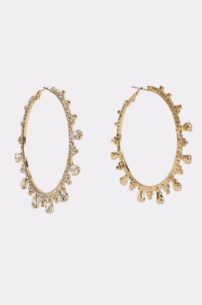 The CRYSTAL DREAMS HOOP EARRINGS are gold oversized hoops adorned with small dangling clear gemstones and encrusted with tiny crystals along the outer edge, beautifully displayed on a plain white background.