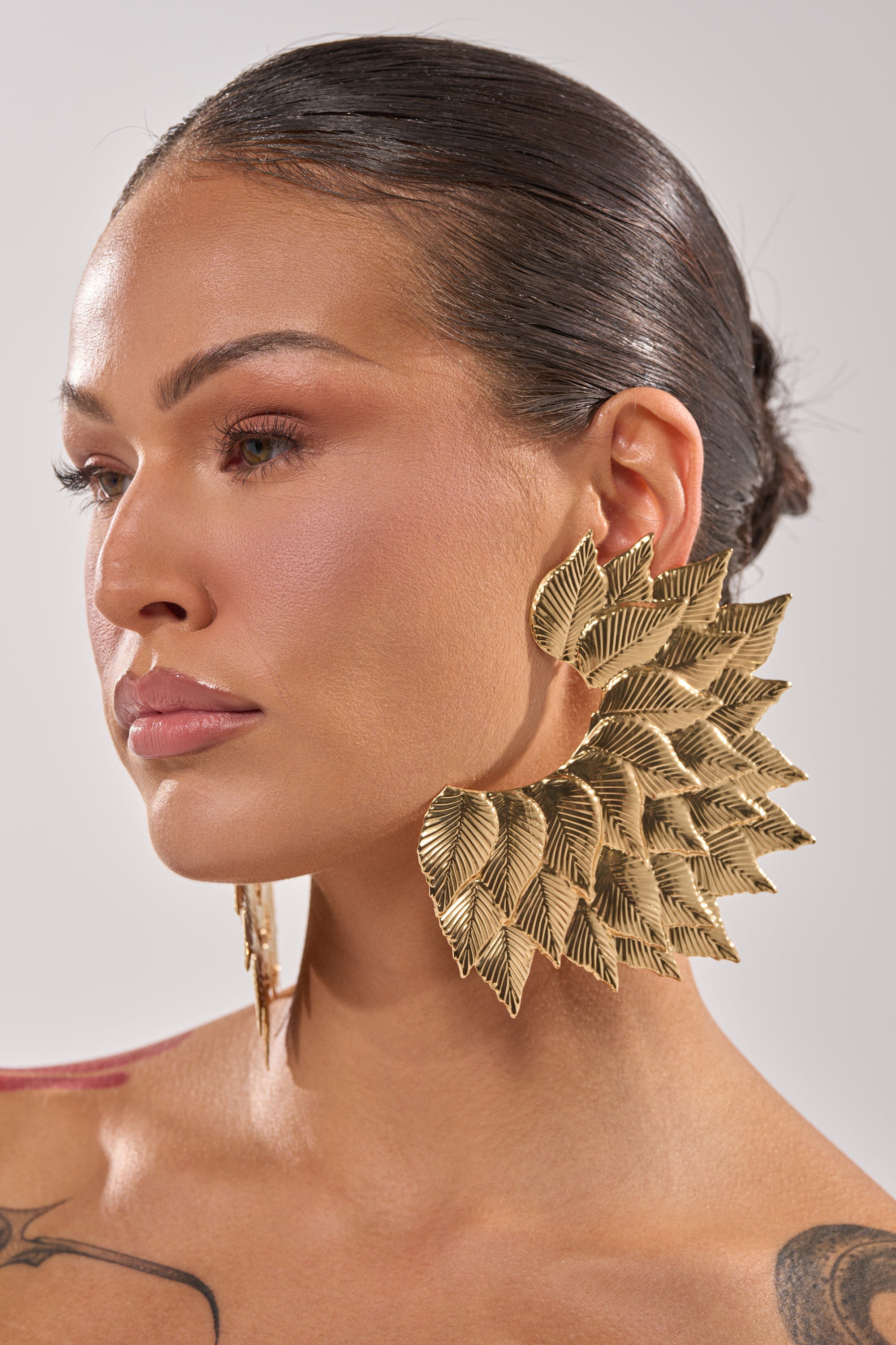 A woman with sleek, pulled-back dark hair wears the SUNSHINE EARRING—oversized gold leaf statement earrings. She has flawless makeup and a shoulder tattoo partly visible against a plain light background. 1