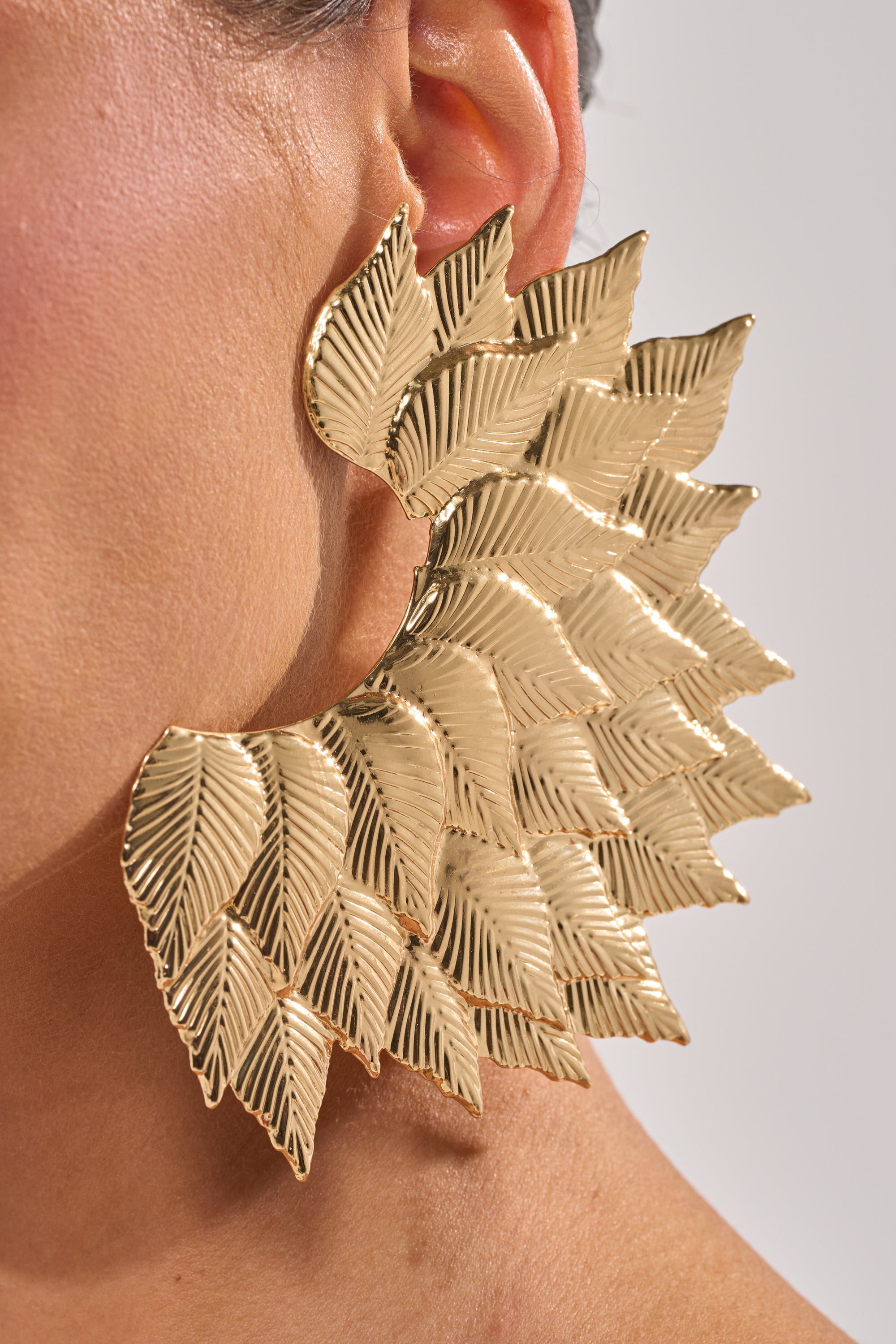 Close-up of a person’s ear adorned with the SUNSHINE EARRING—oversized gold statement earrings with layered, textured leaf shapes that cover much of the ear and extend to part of the cheek. 4