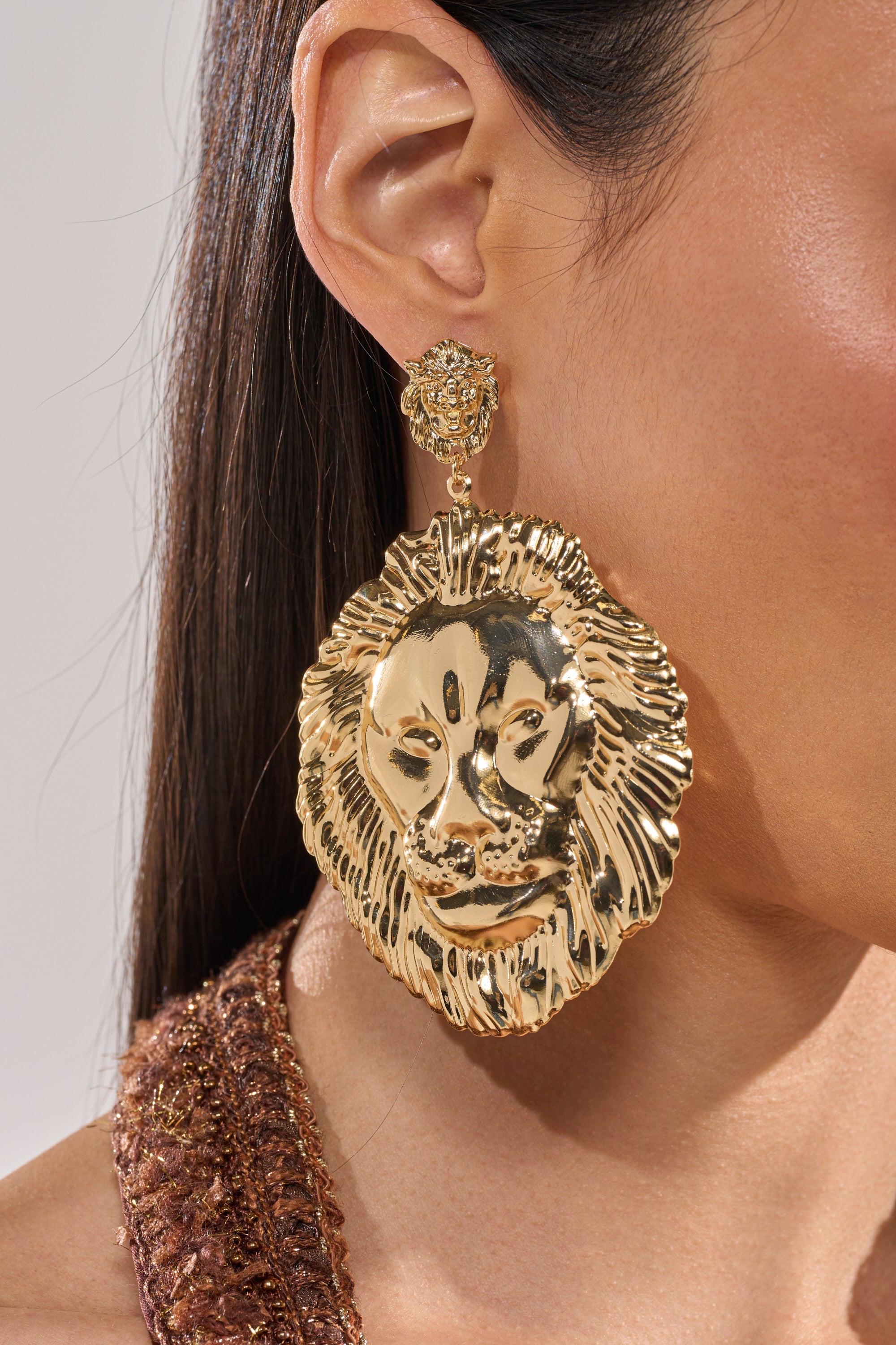 A close-up of a woman’s ear showcases the ROAR EARRING, a striking gold-tone lion-head drop design. Her long, straight hair flows past a textured brown garment. 4
