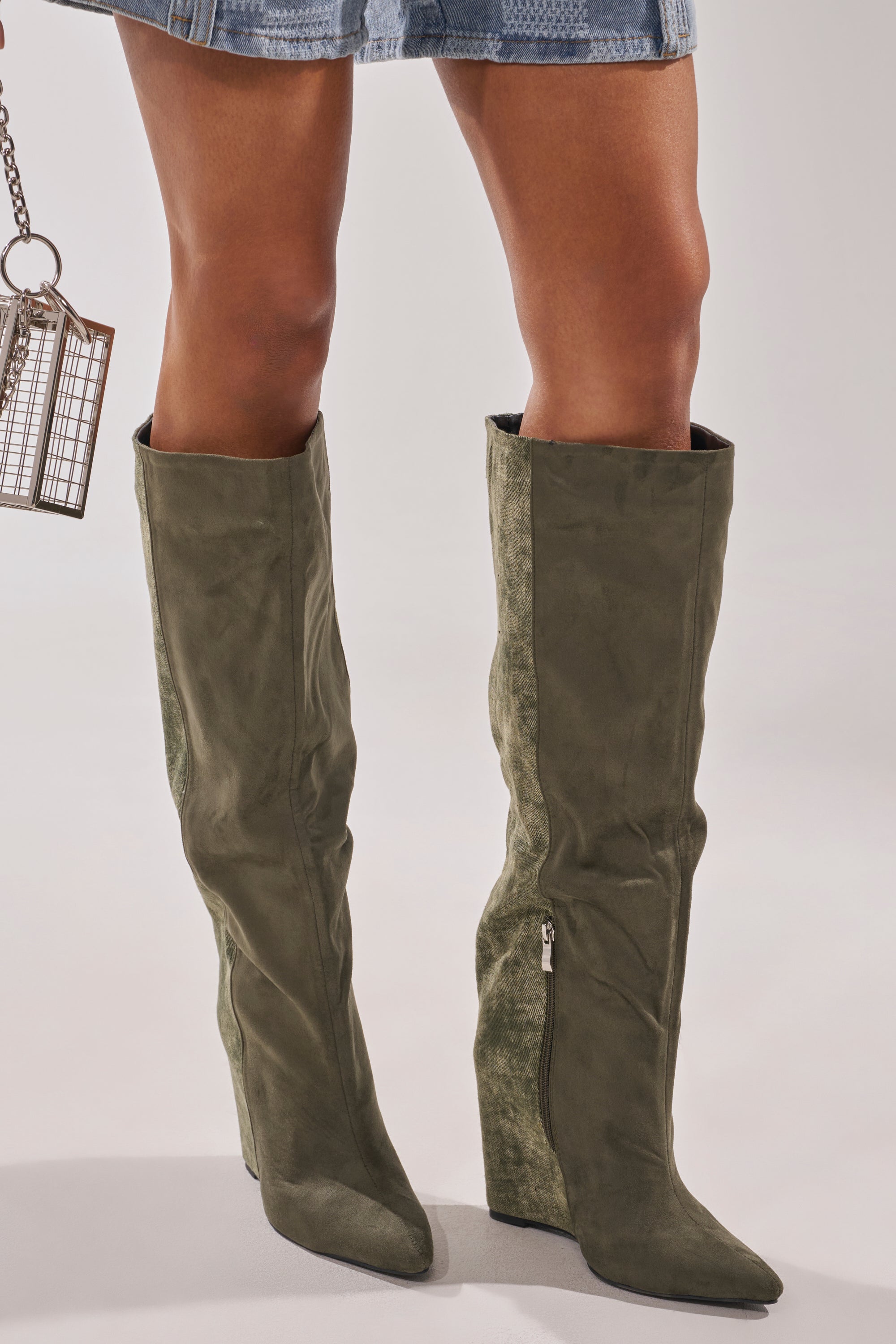 A person wearing the AZALEA WANG GIANNA OLIVE TWO TONED BOOT styled with a short blue denim skirt and holding a small metallic cage-style handbag. 0